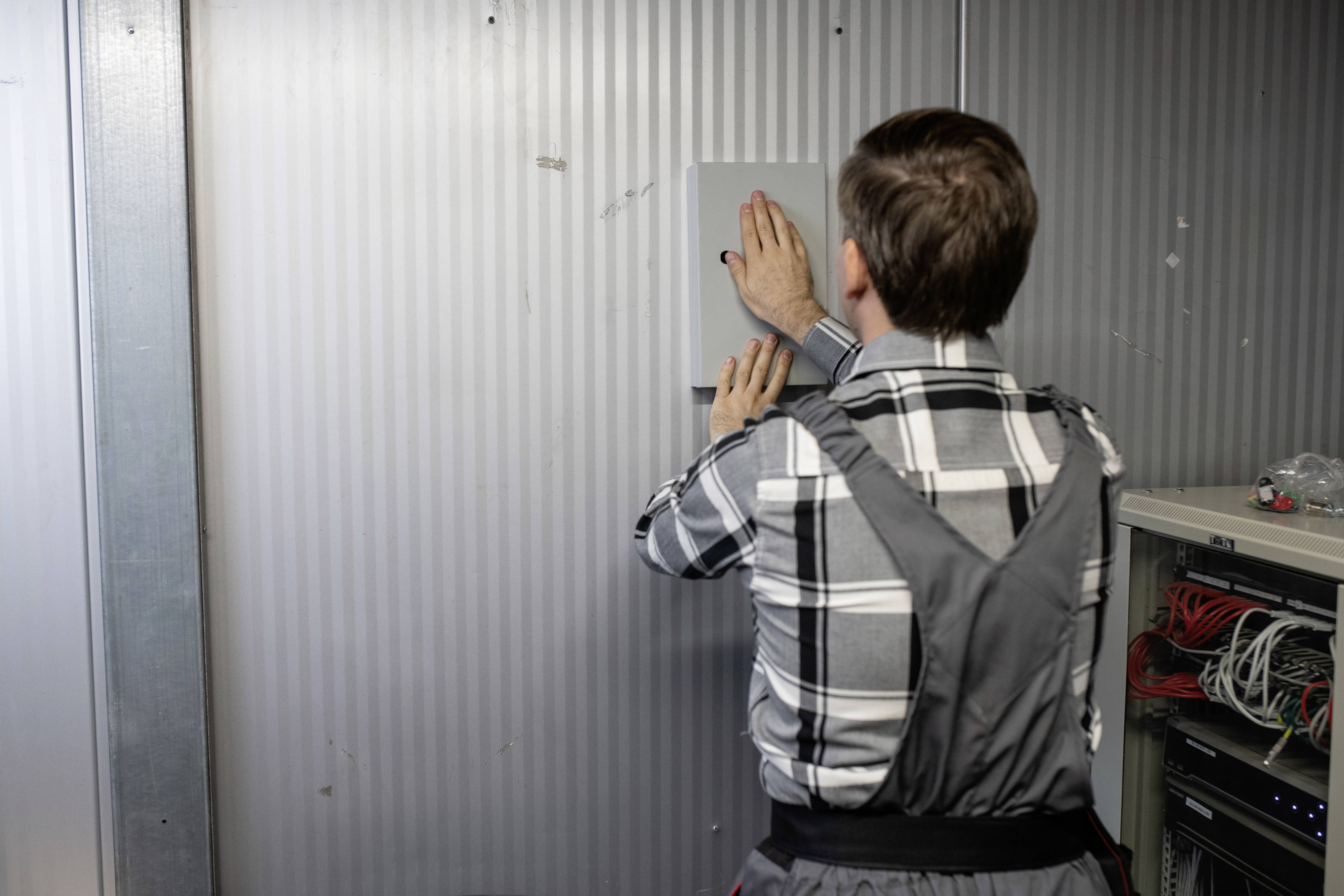Ein Arbeiter in Arbeitskleidung installiert einen Schaltkasten an einer gerippten Metallwand. Rechts sind Kabel sichtbar.