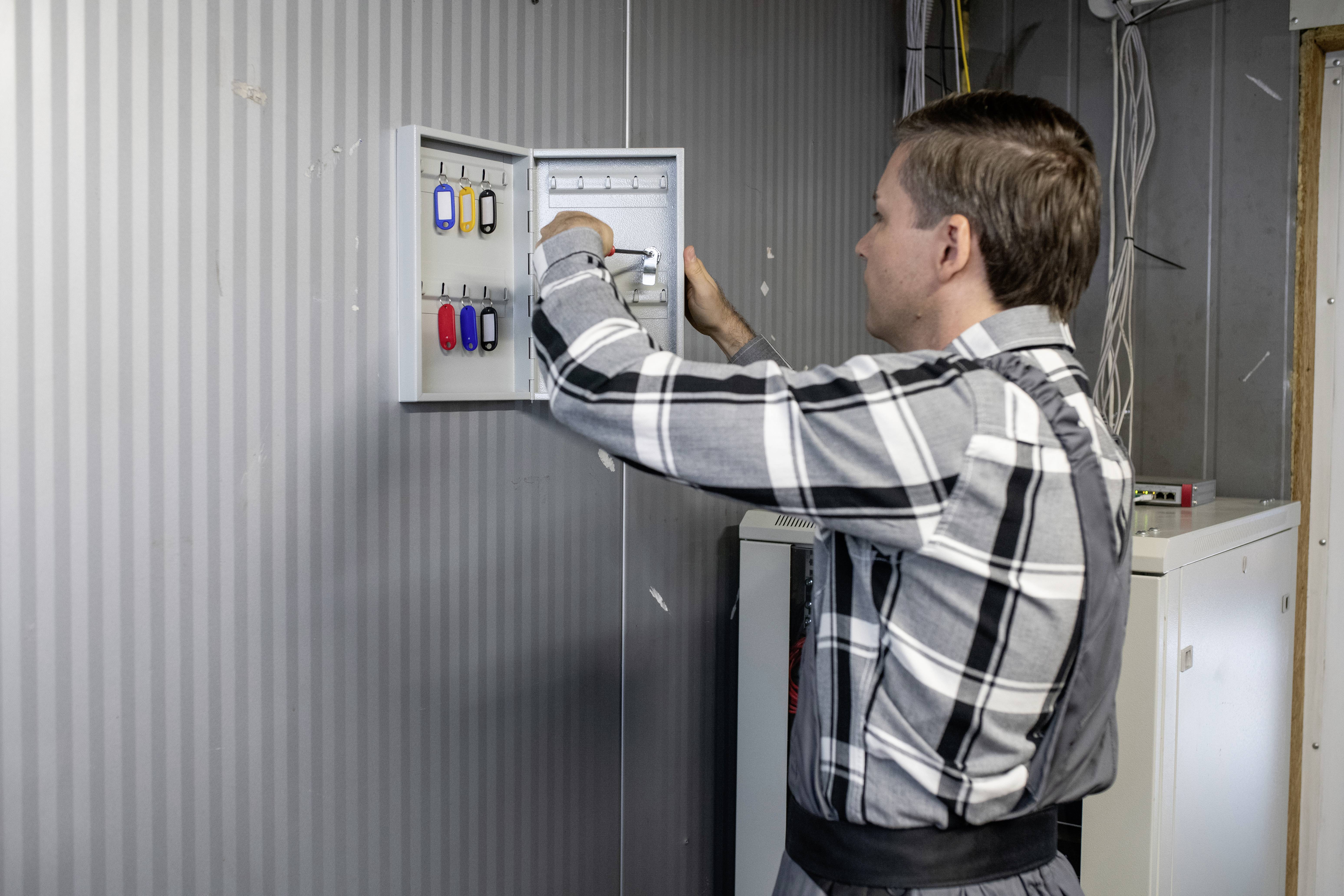 Ein Mann in kariertem Hemd öffnet einen Schlüsselschrank an einer Wand. Im Schrank sind mehrere bunte Schlüsselanhänger sichtbar.