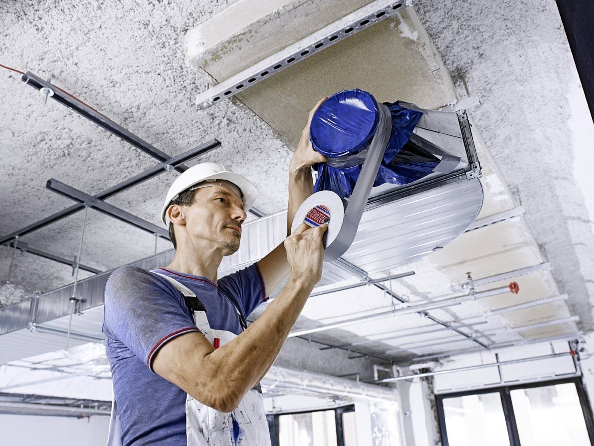 Ein Handwerker mit Helm und Overall dichtet eine Lüftung mit Klebeband in einem unfertigen Raum ab. Baustelle im Innenbereich.