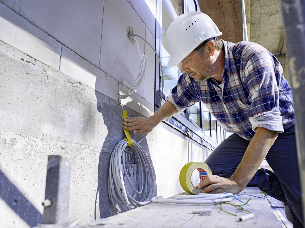 Ein Arbeiter mit Helm installiert elektrische Kabel an einer Außenwand eines Gebäudes. Er hält Klebeband und misst die Kabel.