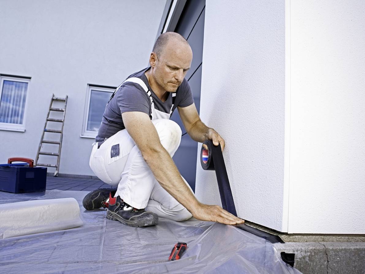 Ein Mann installiert eine Dichtung am Sockel eines Gebäudes. Er trägt Arbeitskleidung und konzentriert sich auf die genaue Platzierung.