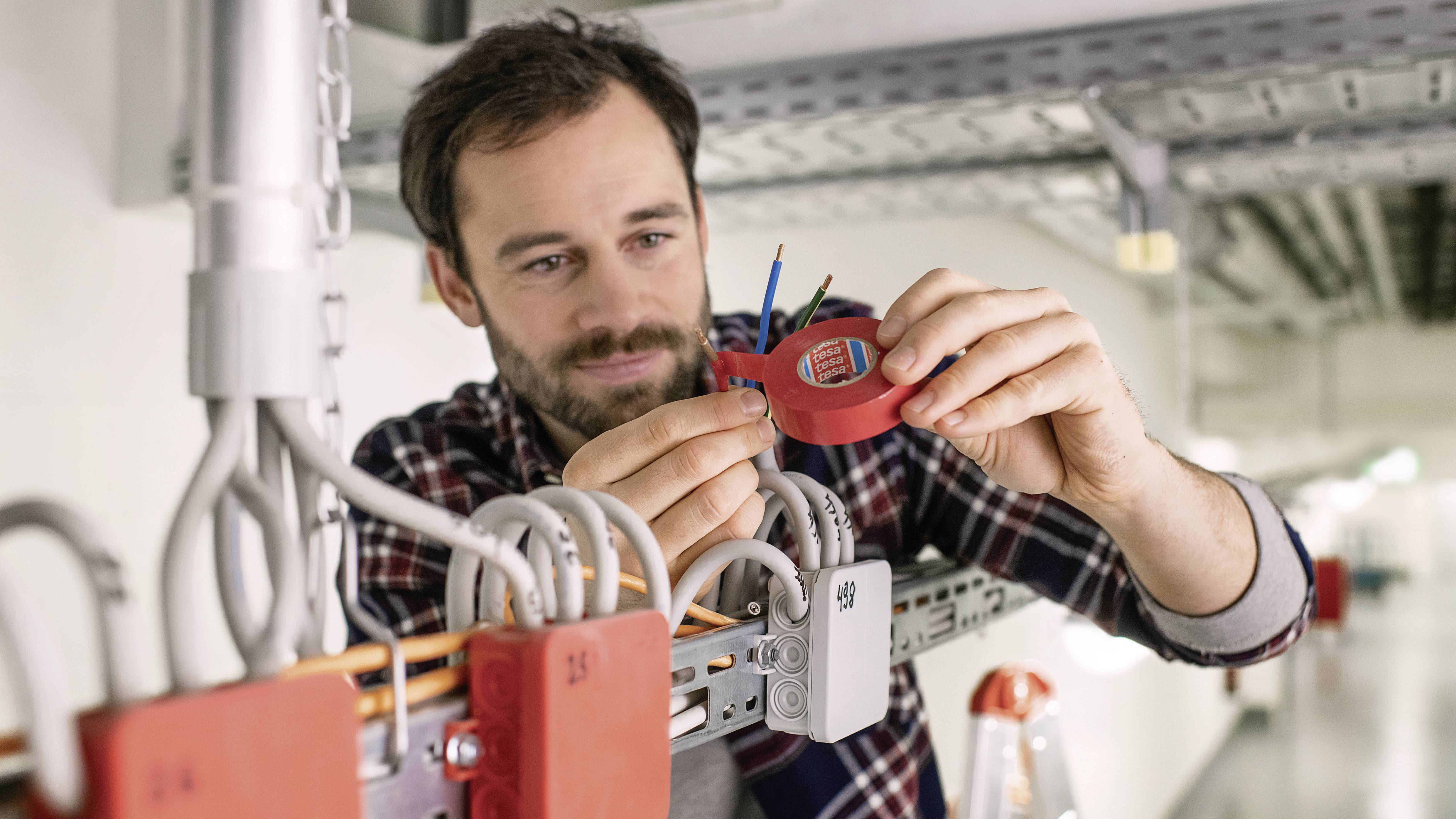 Ein Mann installiert Elektronik in einem Industriegebäude. Er hält Kabel und nutzt ein rotes Gerät, um die Verbindung herzustellen.