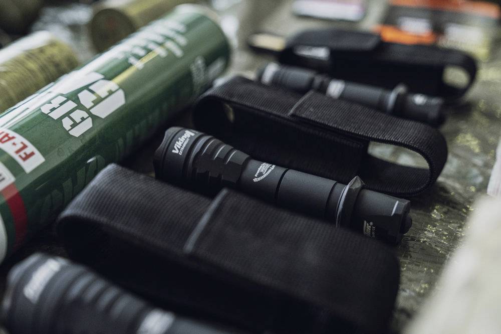 Nahaufnahme mehrerer Taschenlampen in schwarzen Etuis, auf einem Tisch liegend. Im Hintergrund unscharf, ein grüner Zylinder.