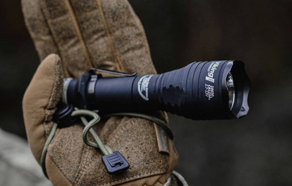 Hand hält schwarze Taschenlampe in Arbeits-Handschuh. Taschenlampe zeigt nach vorne, Schnur umwickelt Griff, Text nicht lesbar.