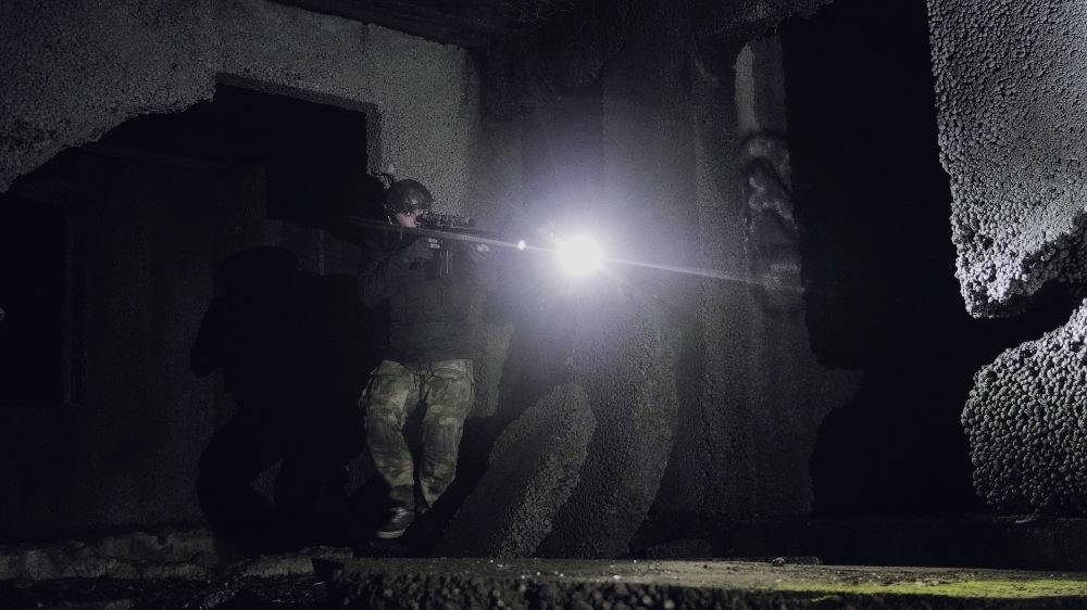 Eine Person im Tarnanzug steht in einem dunklen, verfallenen Gebäude und leuchtet mit einer Taschenlampe auf etwas im Vordergrund.
