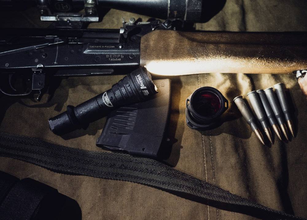 Eine Waffe mit Holzschaft liegt auf einem Tisch, umgeben von einer Taschenlampe, einem Magazin, einem Objektiv und fünf Patronen.