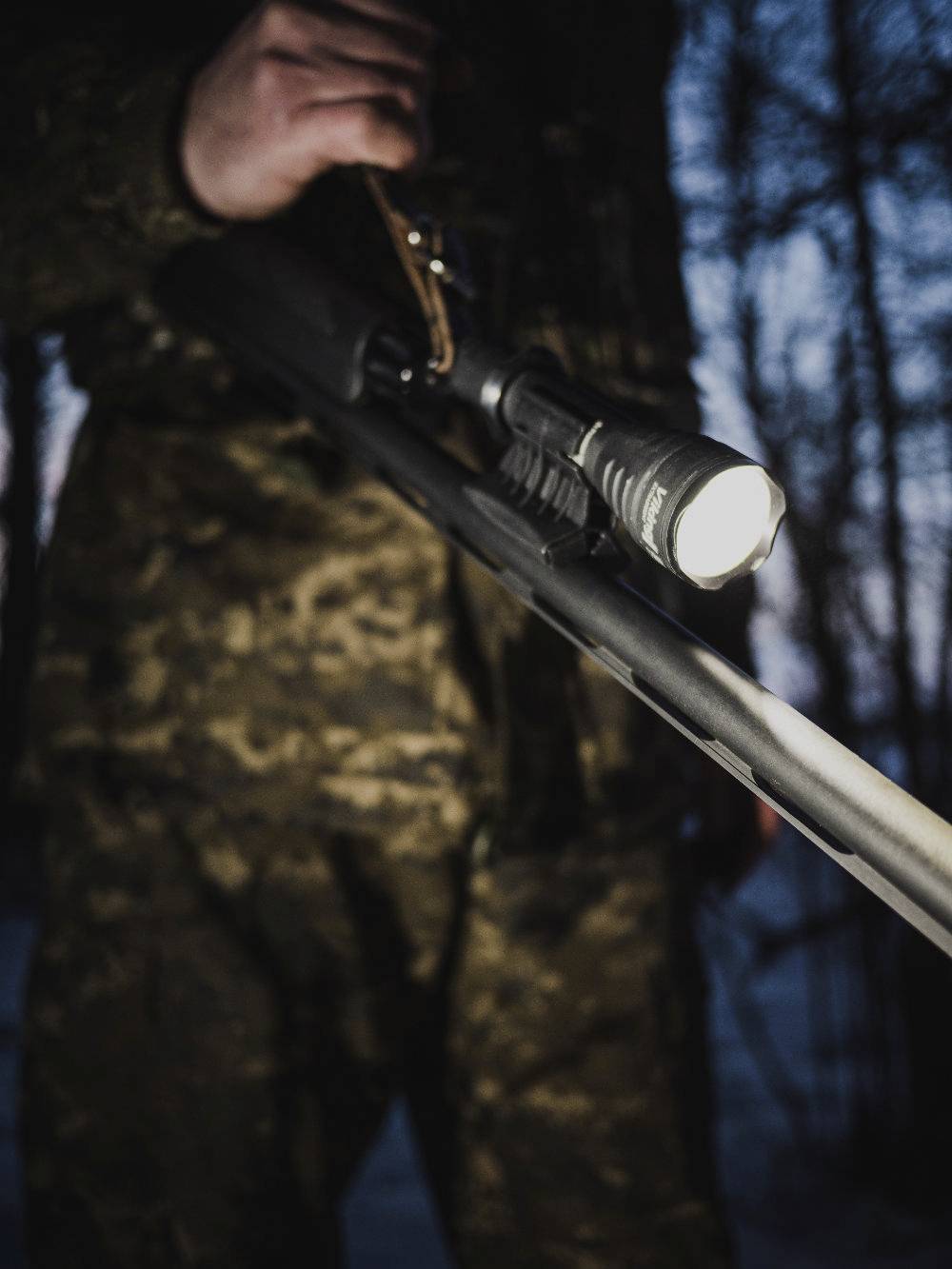 Person in Tarnkleidung hält ein Gewehr mit montierter Taschenlampe, im Hintergrund unscharfe Bäume bei Dämmerung.