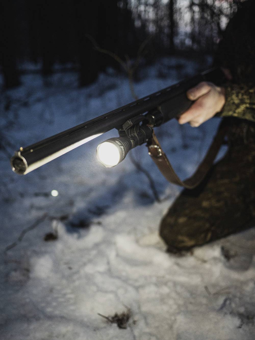 Person kniet im Schnee, hält ein Gewehr mit Taschenlampe. Die Umgebung wirkt dunkel und winterlich, vermutlich im Wald.