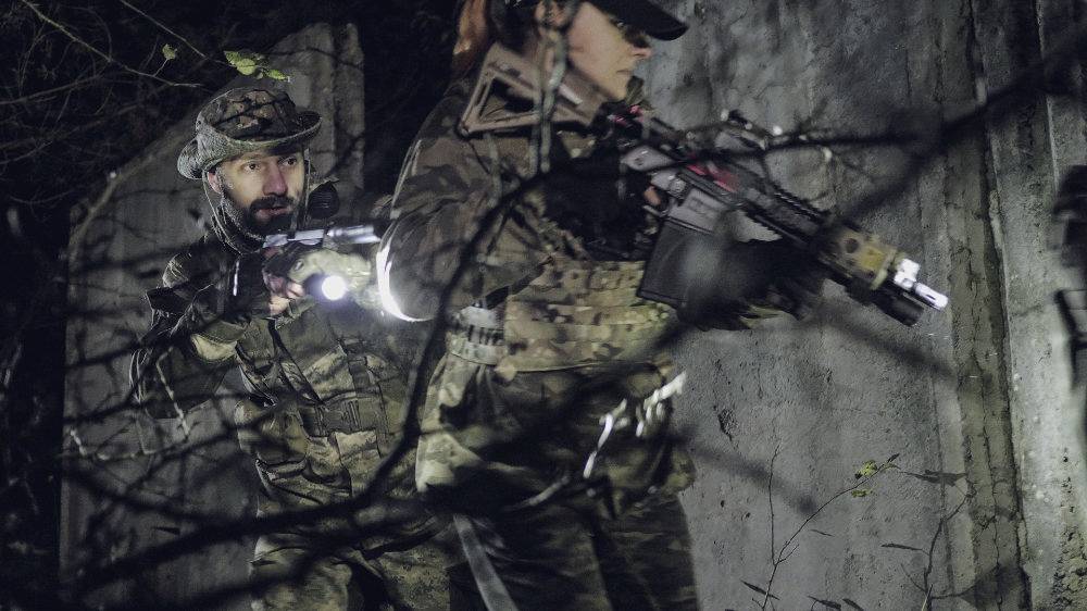 Zwei Personen in Tarnkleidung mit Gewehren und Taschenlampen durchsuchen eine dunkle Umgebung, wie ein taktisches Manöver.