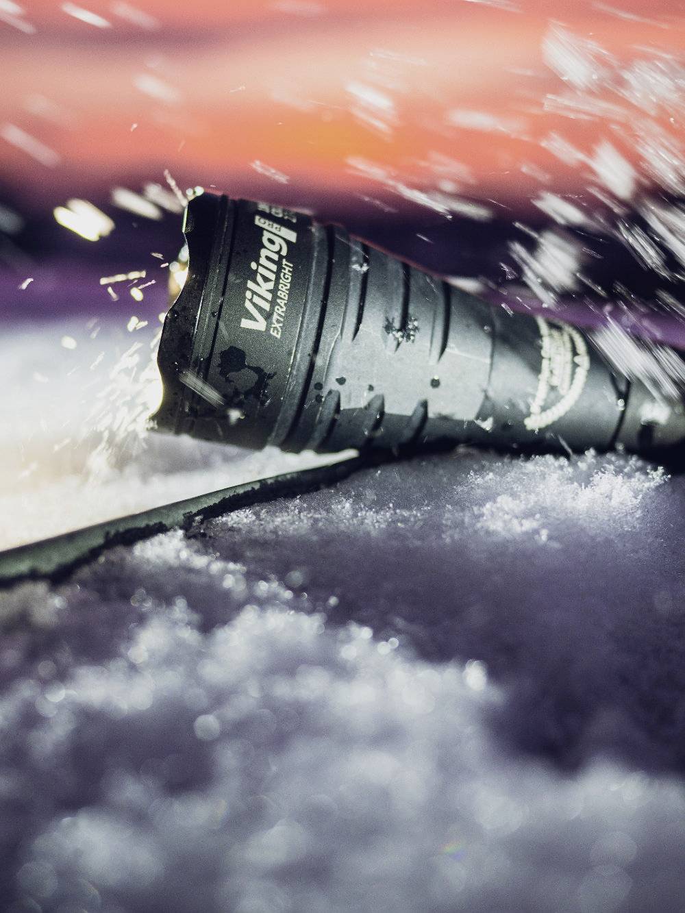 Taschenlampe liegt in Schnee, umgeben von spritzendem Wasser. Hintergrund ist unscharf und zeigt lebendige Rottöne.