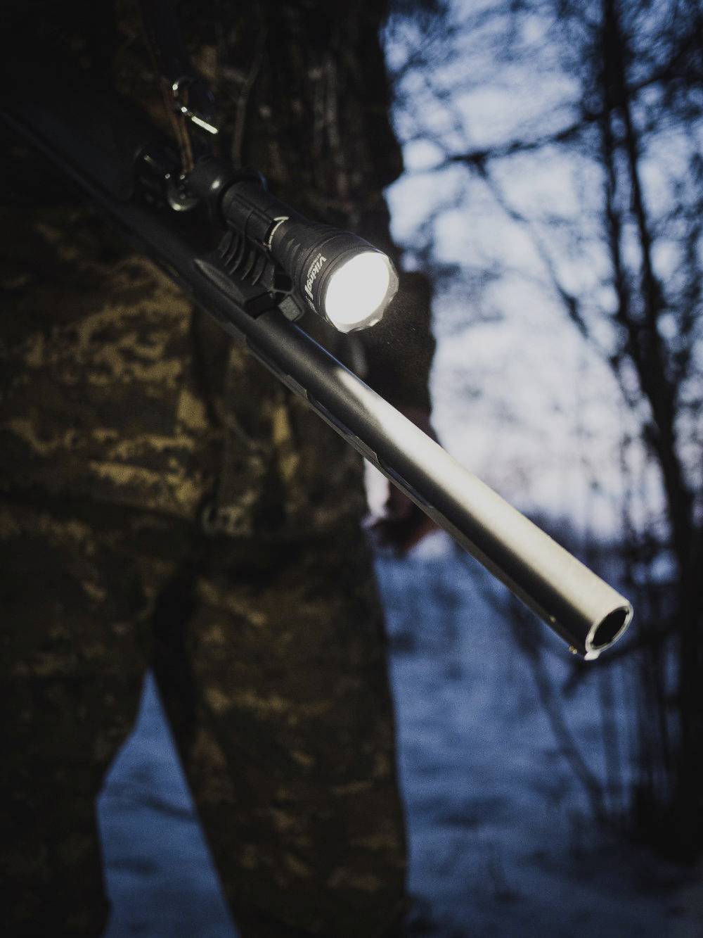 Eine Person in Tarnkleidung hält eine Gewehr mit Taschenlampe im Wald bei Dämmerung.