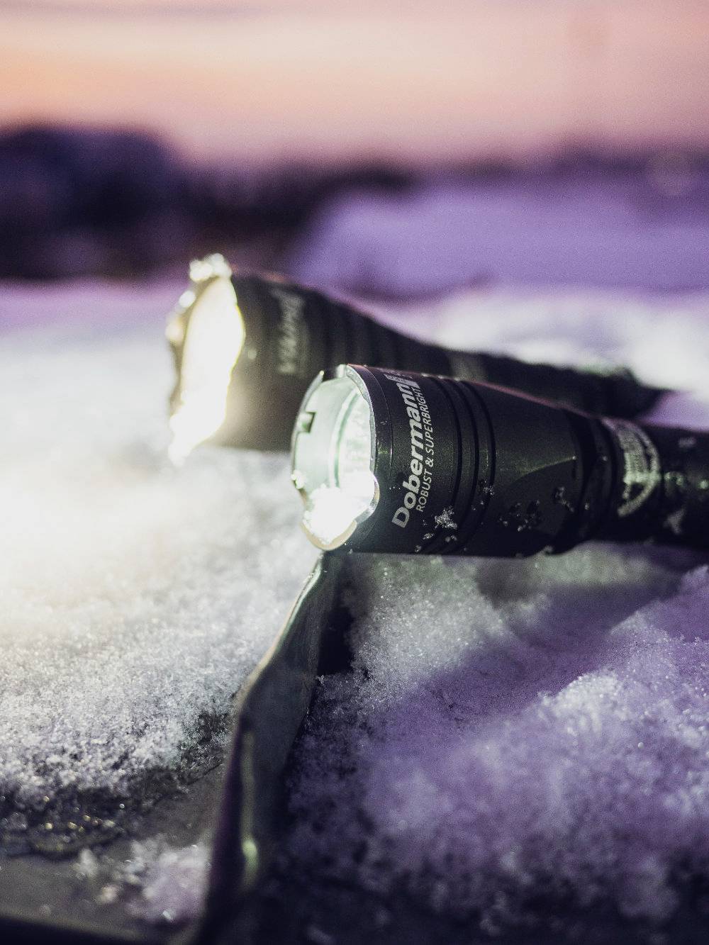Zwei Taschenlampen leuchten im Schnee bei Sonnenuntergang. Die vordere ist eingeschaltet und strahlt hell auf die kristalline Oberfläche.