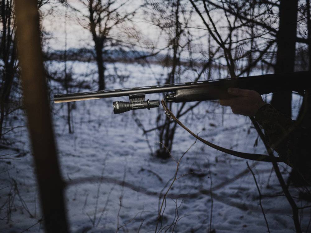 Eine Person hält ein Gewehr im verschneiten Wald bei Dämmerung. Bäume und Äste umgeben die Szene, die eine ruhige Atmosphäre vermittelt.