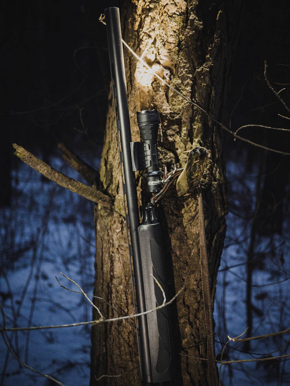 Ein Gewehr lehnt gegen einen Baumstamm in einem verschneiten Wald bei Nacht.