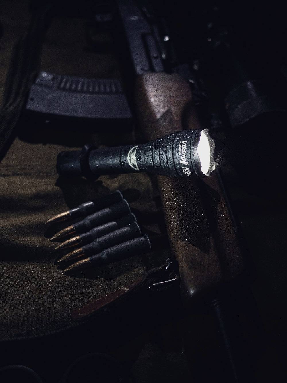 Taschenlampe leuchtet auf ein Gewehr und Patronen, die auf einem Tuch liegen. Die Szene ist dunkel, fokussiert auf die Waffen.