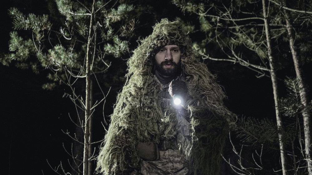 Person, in Tarnkleidung und mit Taschenlampe, steht nachts in einem Wald.
