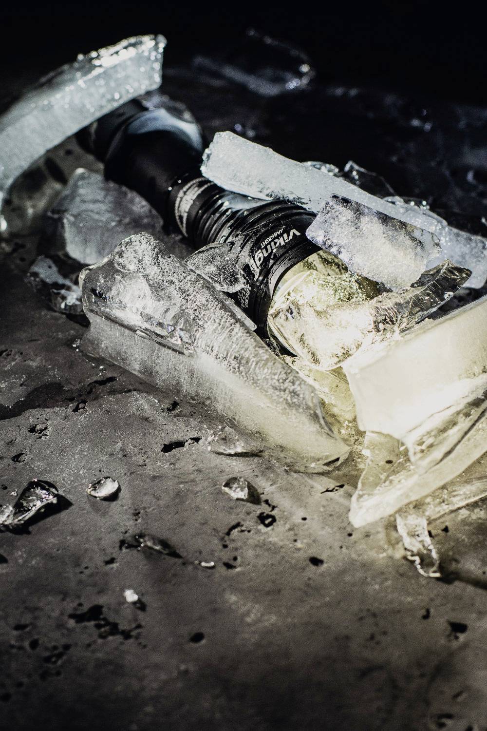 Eine zerbrochene Glühbirne liegt auf einem dunklen, eisbedeckten Untergrund. Die Eisstücke sind ungleichmäßig um die Birne verteilt.