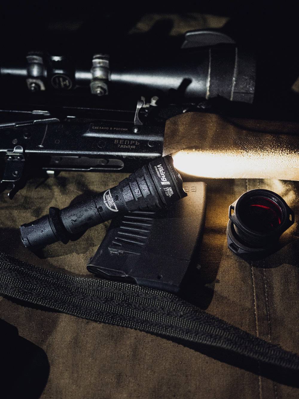 Ein Gewehr mit angebrachtem Zielfernrohr, eine Taschenlampe, ein Magazin und ein Objektivaufsatz liegen zusammen auf braunem Stoff.