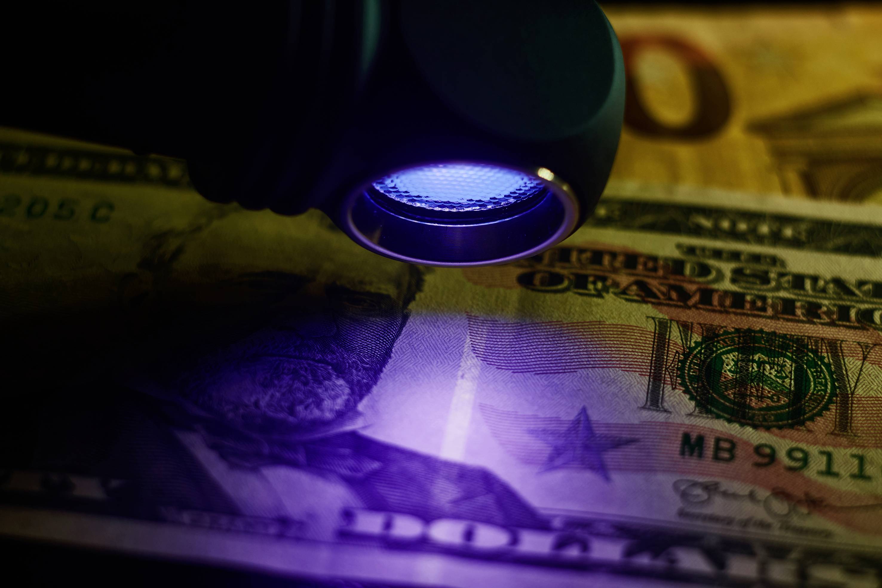 Eine UV-Lampe beleuchtet einen US-Dollar-Schein. Sicherheitsmerkmale sind sichtbar. Im Hintergrund ein weiterer Schein.