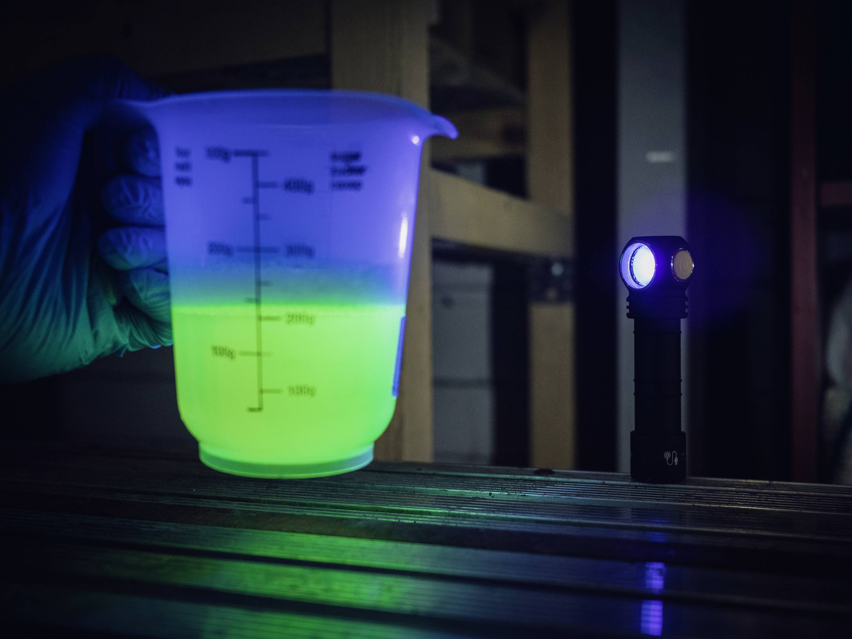 Eine Hand in einem Handschuh hält einen Becher mit fluoreszierender grüner Flüssigkeit, beleuchtet von einer kleinen Taschenlampe.
