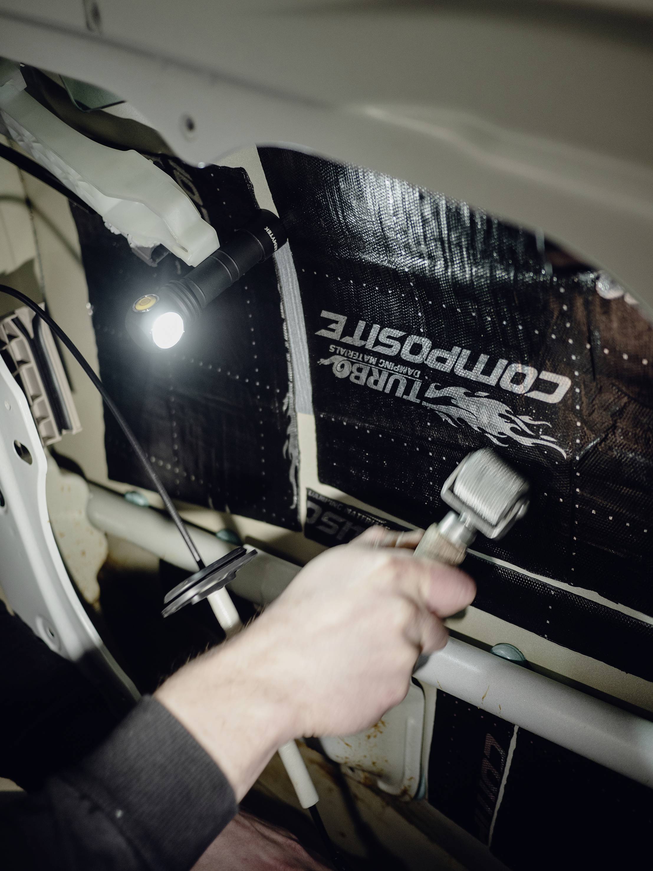 Eine Person im Auto arbeitet mit einem Werkzeug an der Dämmung im Innenraum, beleuchtet von einer Lampe.