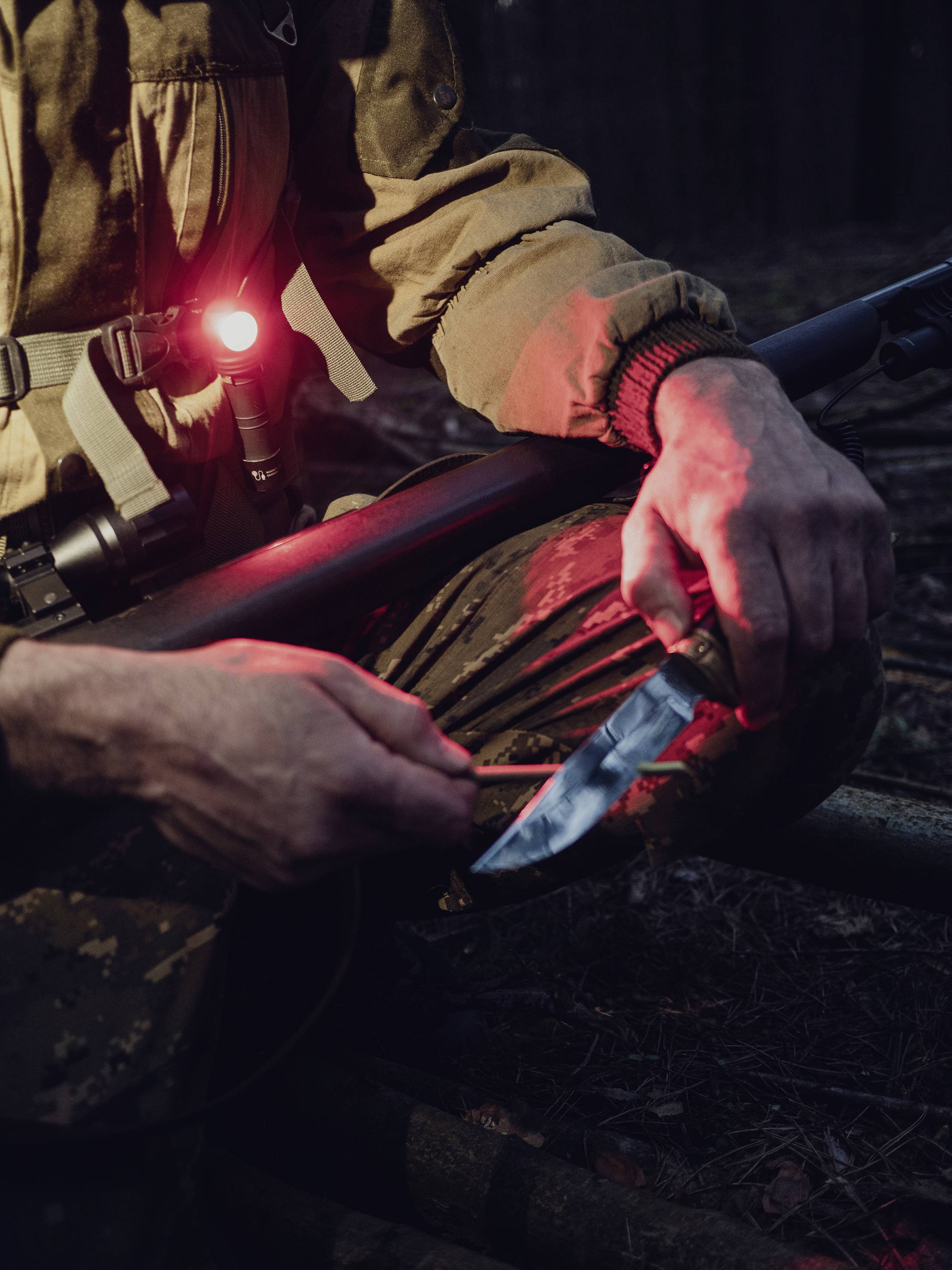 Person im Wald hält ein Gewehr und schärft ein Messer mit Holz. Rotes Licht beleuchtet die Szene. Atmosphäre ist dunkel und konzentriert.