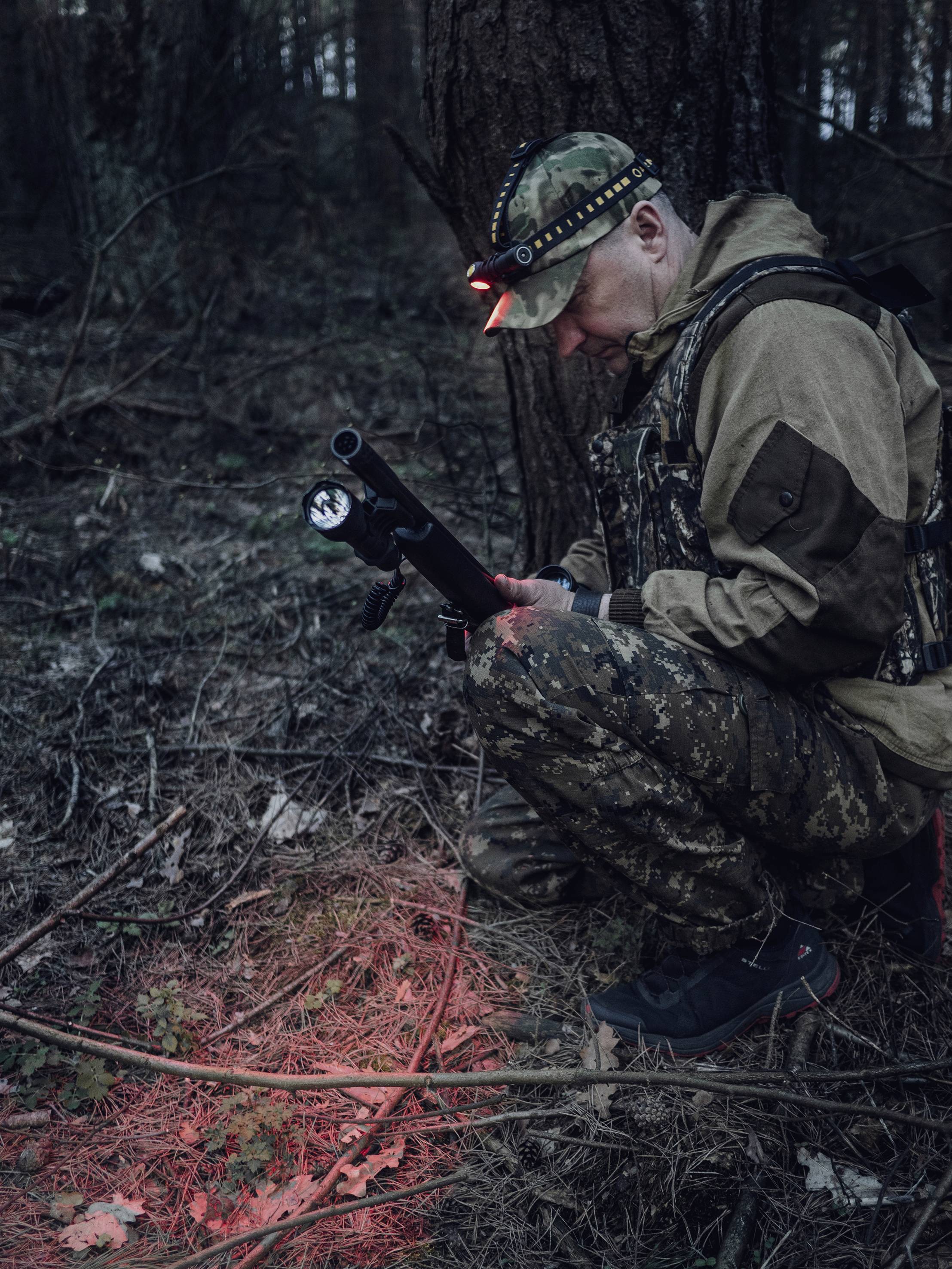 Jäger in Tarnkleidung kniet im Wald, untersucht Boden mit Taschenlampe. Rotes Licht beleuchtet Laub.