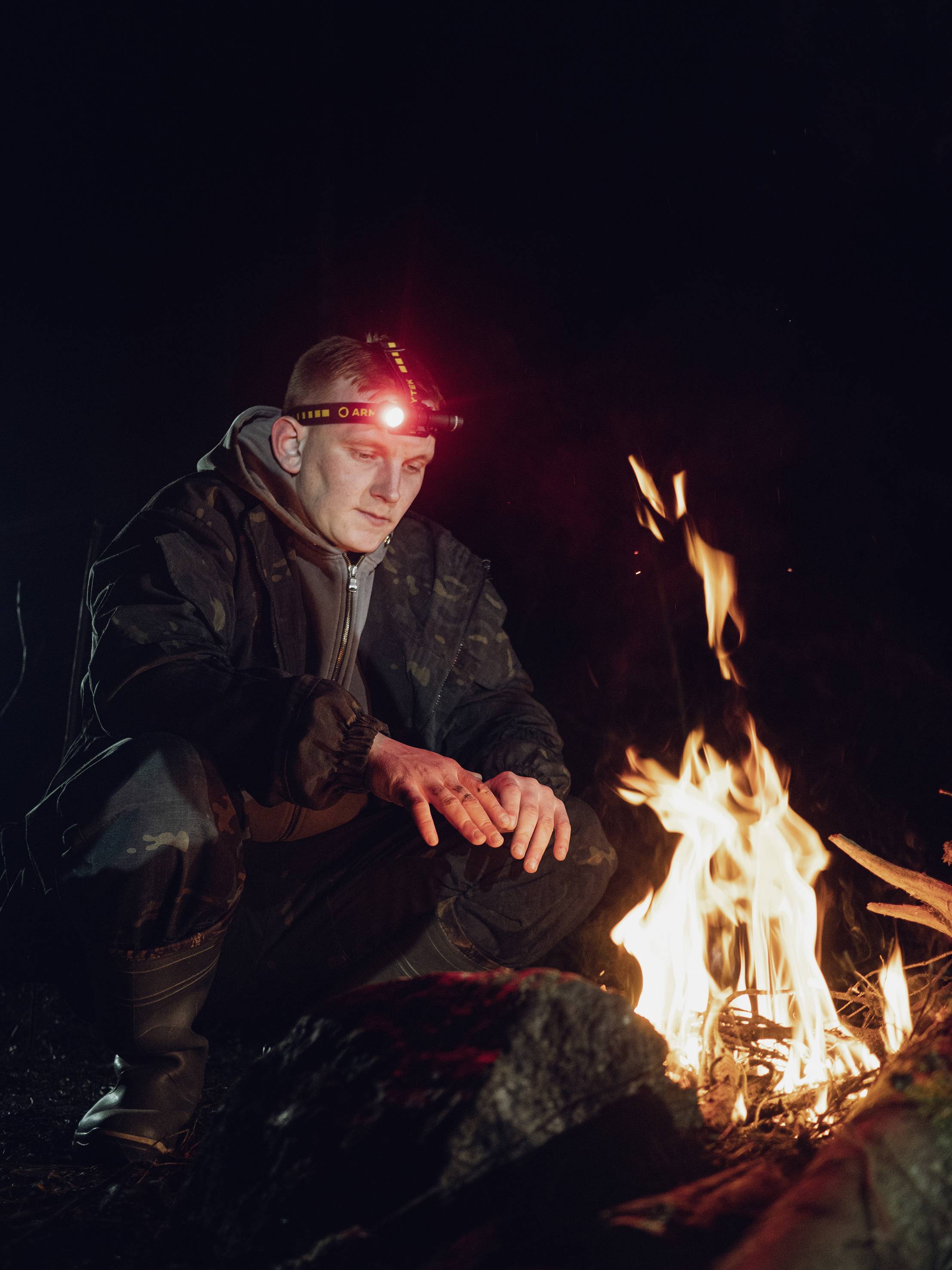 Eine Person mit Stirnlampe sitzt nachts im Freien am Lagerfeuer. Sie wärmt ihre Hände an den Flammen.