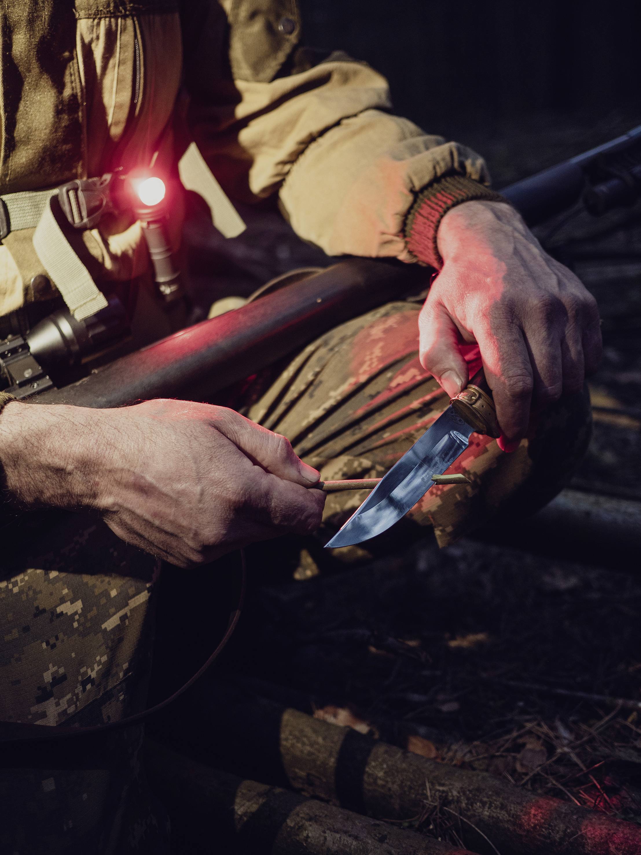 Eine Person in Tarnkleidung hält und schärft ein Messer. Ein rotes Licht leuchtet an der Kleidung. Die Umgebung ist dunkel.