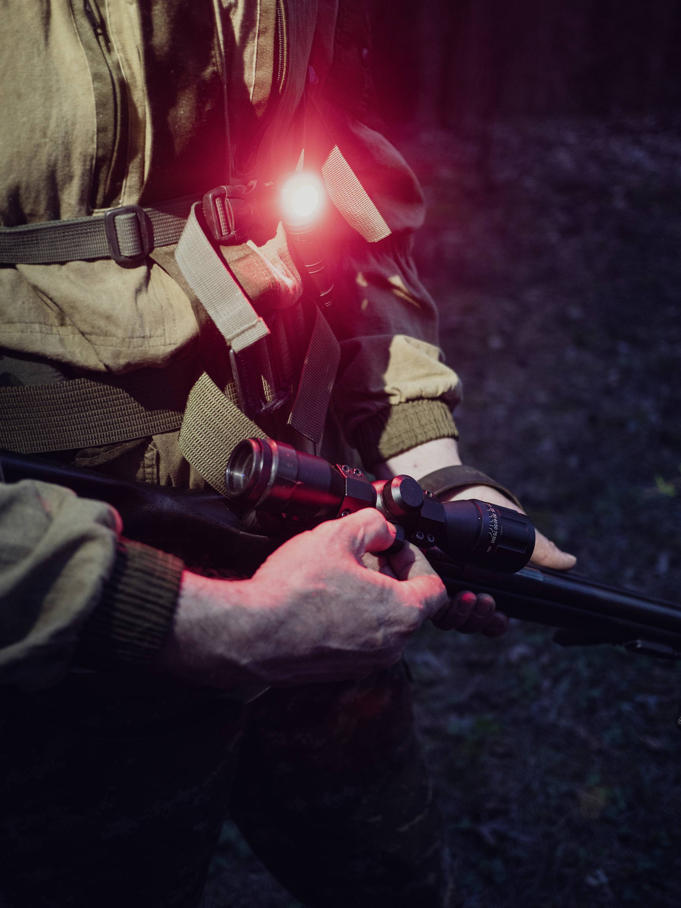 Eine Person in Tarnkleidung hält ein Gewehr mit einem Zielfernrohr. Ein rotes Licht leuchtet von der Brusttasche.