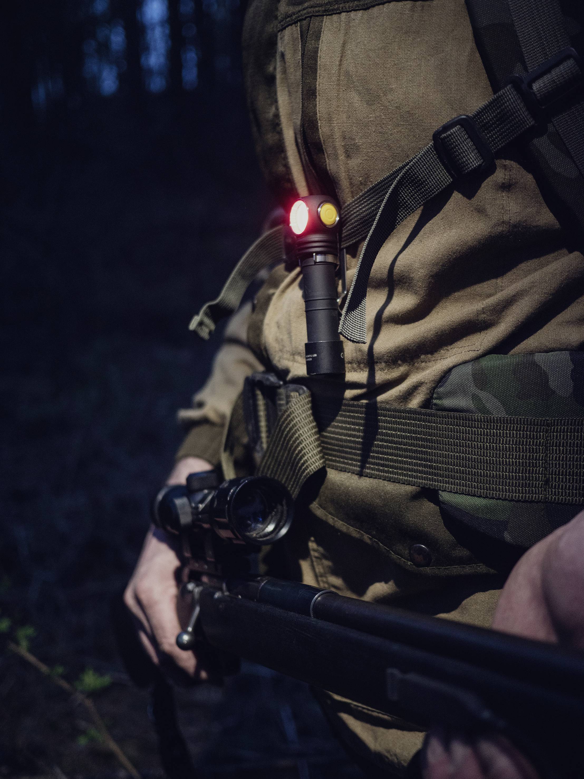 Person in Tarnkleidung hält ein Gewehr bei Dämmerlicht, an der Brust befestigt eine Taschenlampe mit rotem und gelbem Licht.
