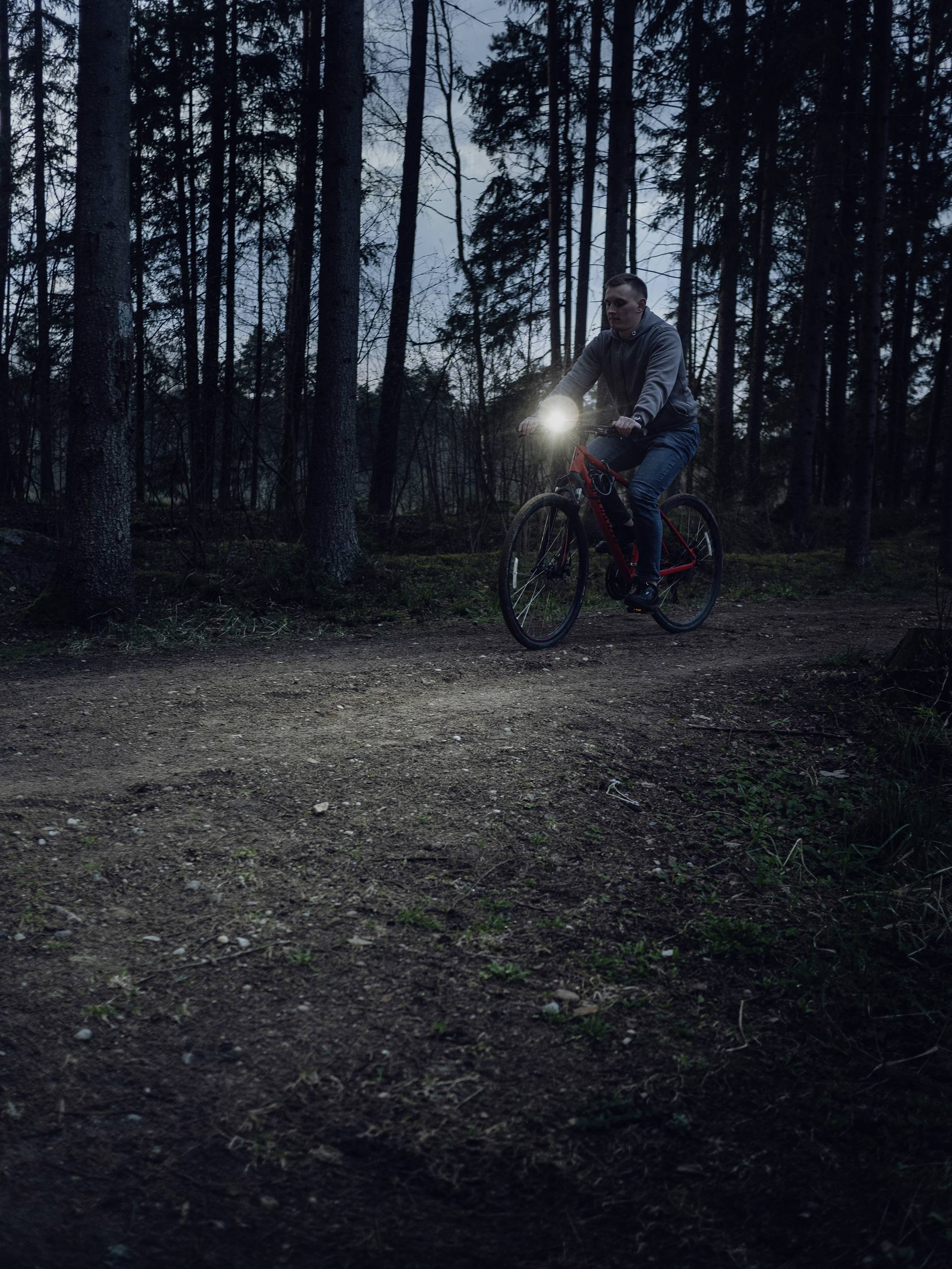Eine Person fährt in der Dämmerung mit einem Fahrrad auf einem Waldweg. Ein Scheinwerfer beleuchtet den Weg vor ihnen.