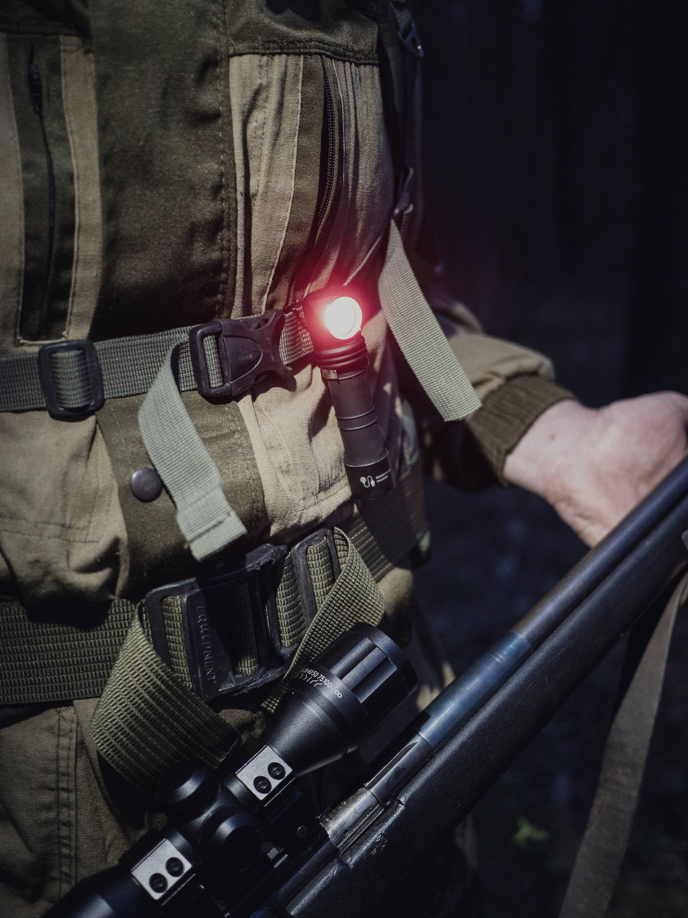 Person in dunkler Kleidung hält ein Gewehr im Wald, mit rotem Licht an der Ausrüstung.