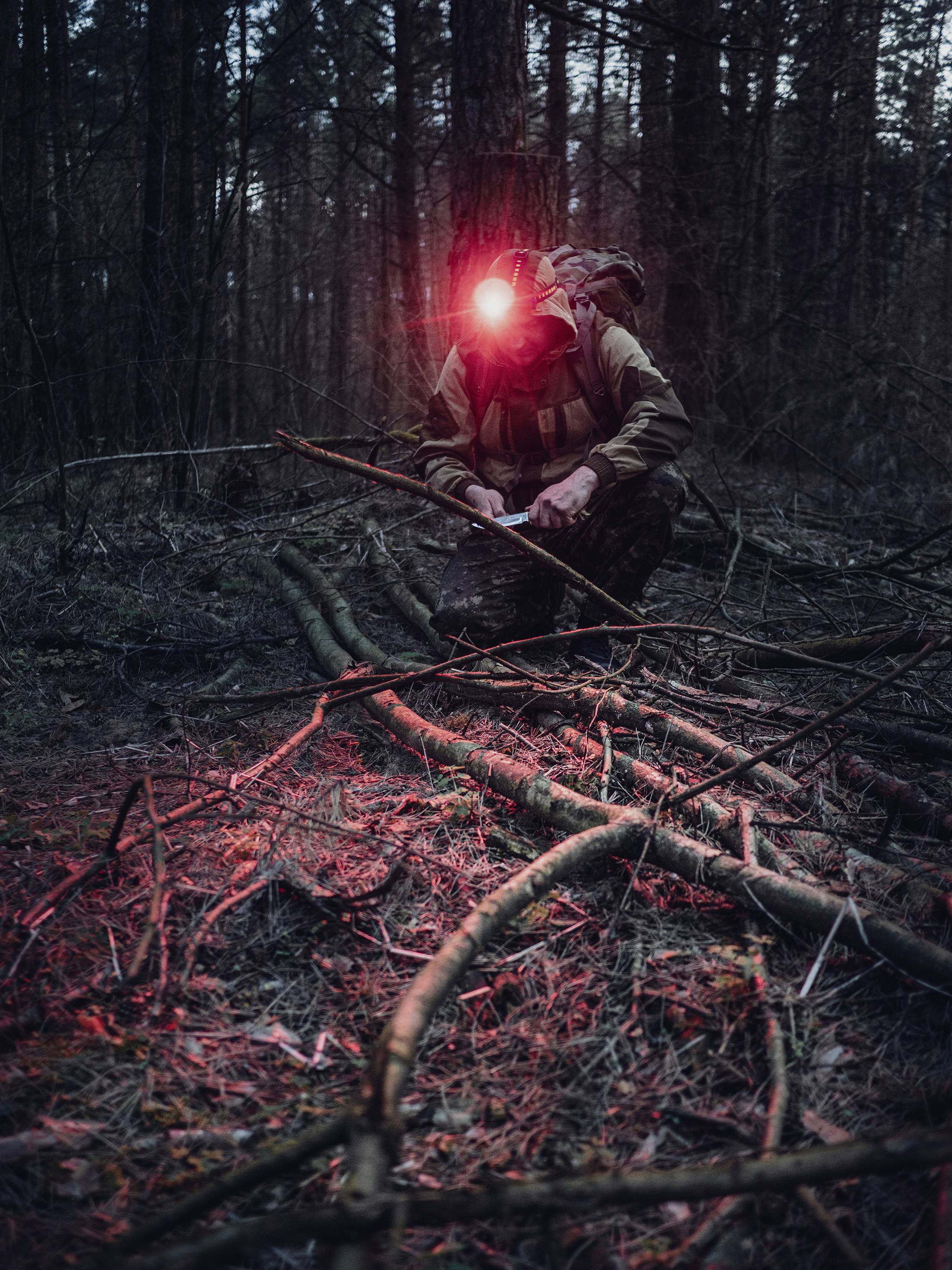 Eine Person im dunklen Wald, kniend über gesammelten Zweigen. Sie trägt einen Rucksack und eine leuchtende Stirnlampe, die die Szene beleuchtet.