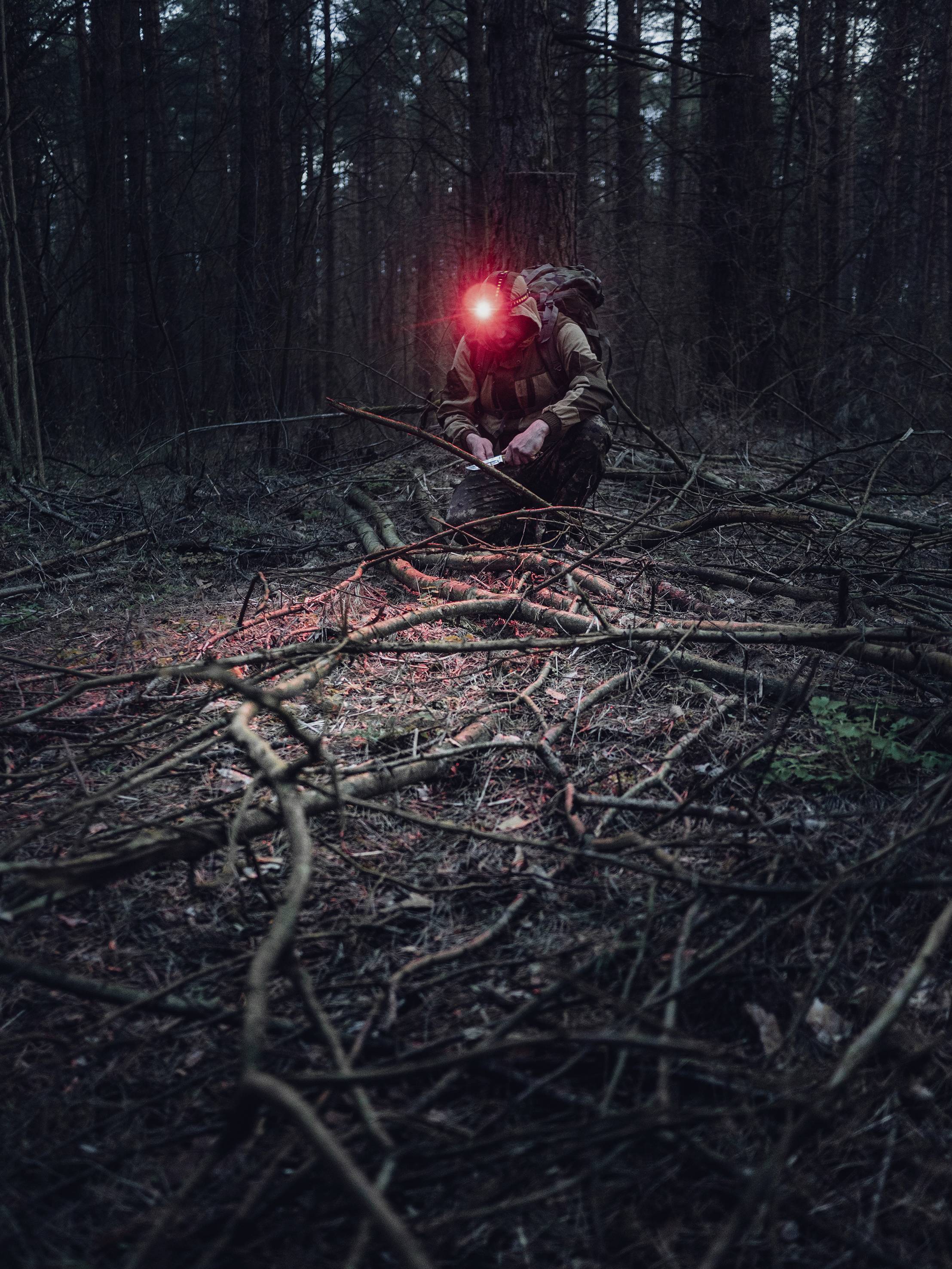 Eine Person kniet in einem Wald und trägt ein rotes Stirnlicht. Es ist dunkel, und das Licht beleuchtet einige Zweige auf dem Boden.