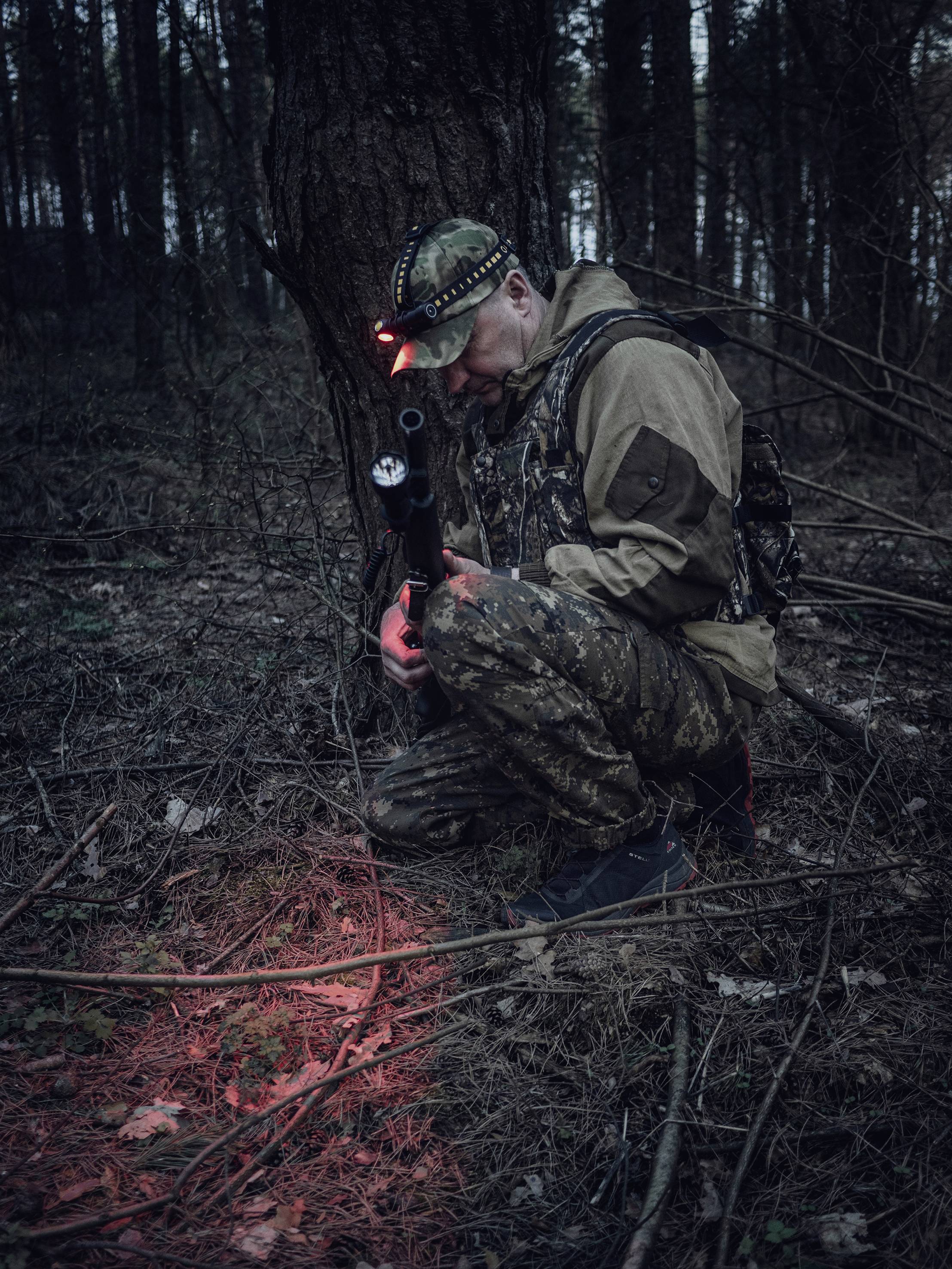 Person in Tarnkleidung kniet im Wald mit rotem Licht. Gewehrauflage, konzentriert auf den Boden. Jagd- oder Nachtaktivität.
