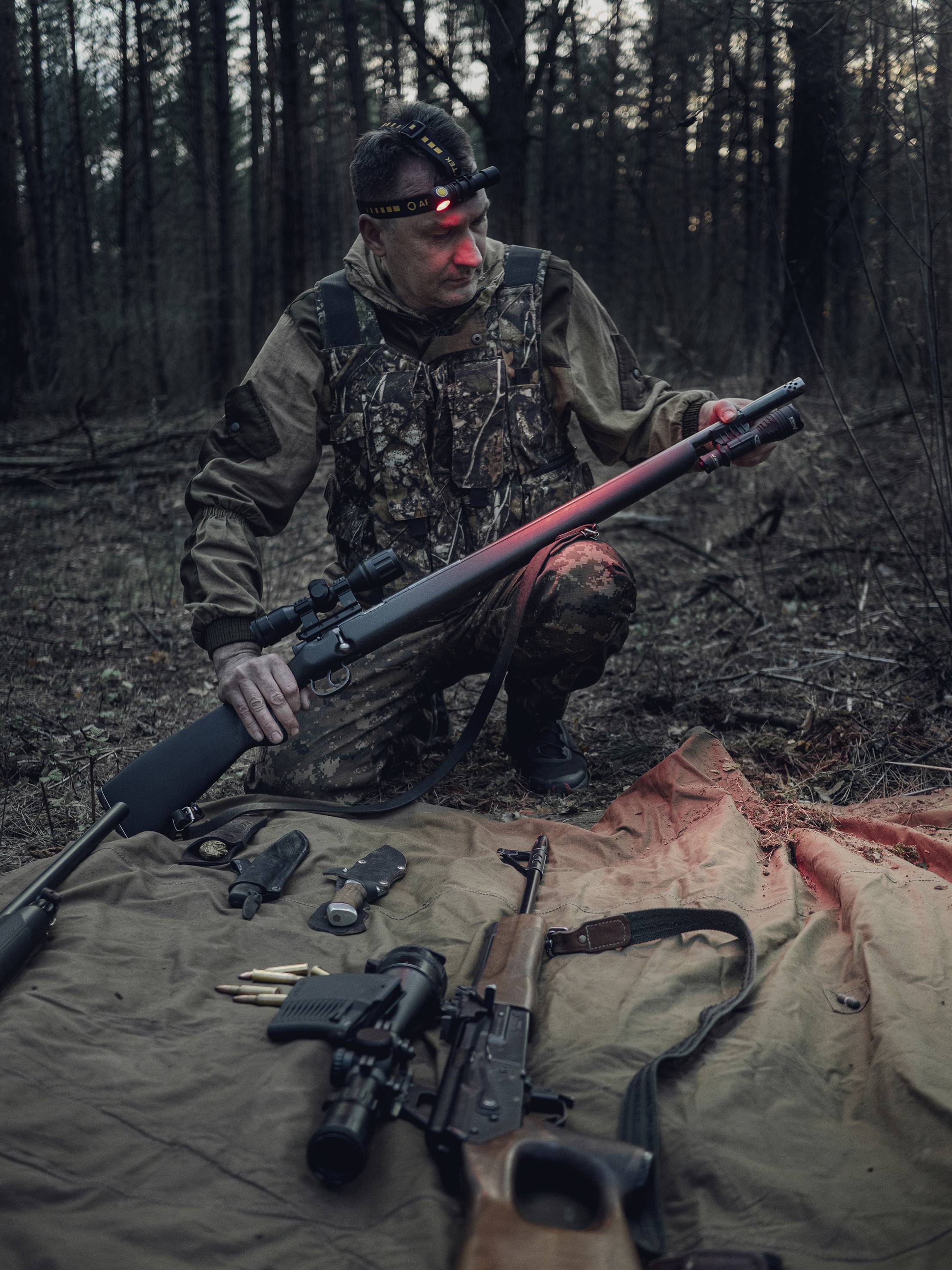 Ein Jäger im Wald sitzt auf einem knie und hält ein Gewehr. Vor ihm liegen zwei Gewehre und Munition auf einer Plane. Dämmerung im Hintergrund.