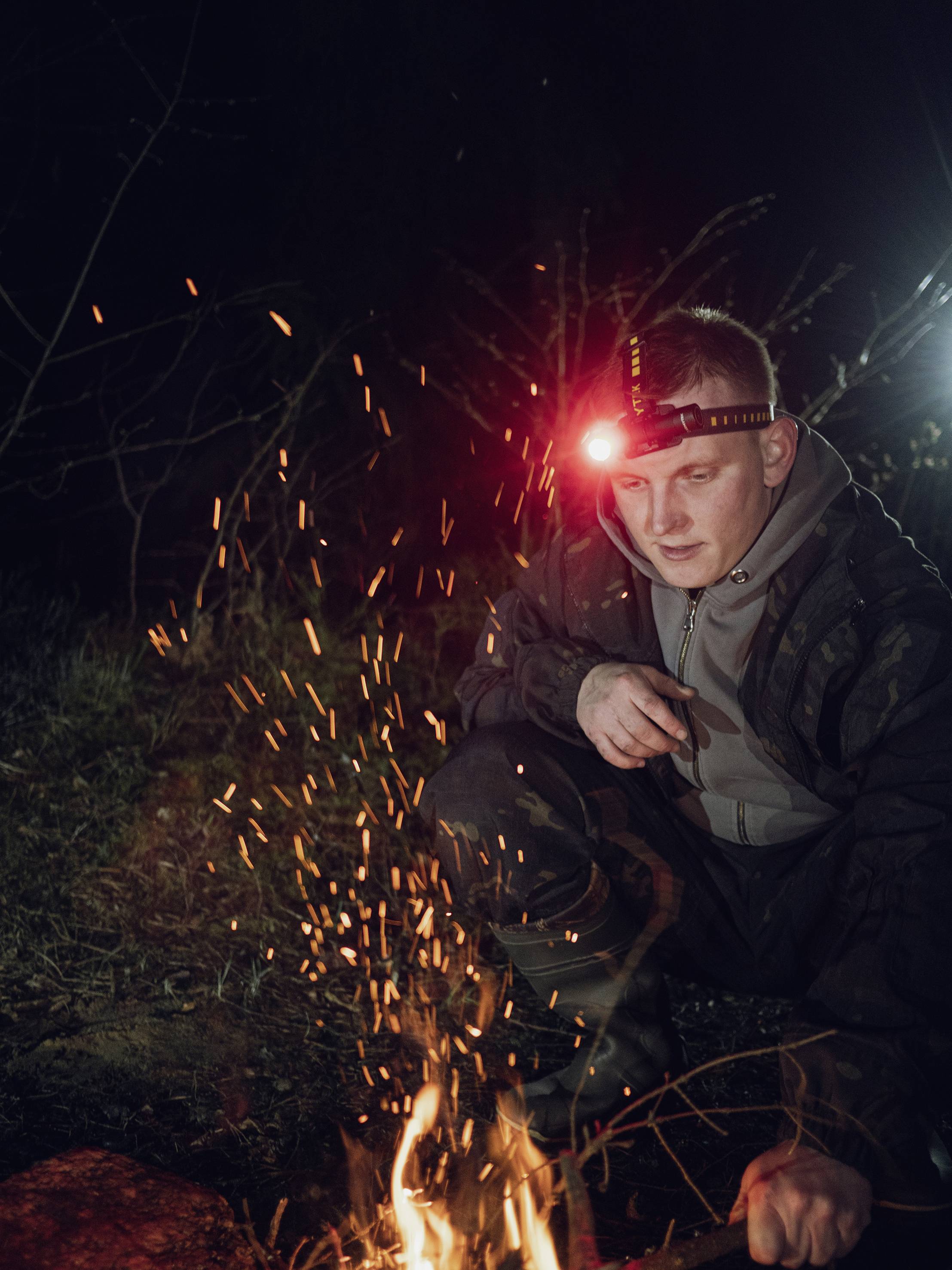 Eine Person mit Stirnlampe kniet im Dunkeln und beobachtet ein kleines Lagerfeuer, aus dem Funken sprühen.