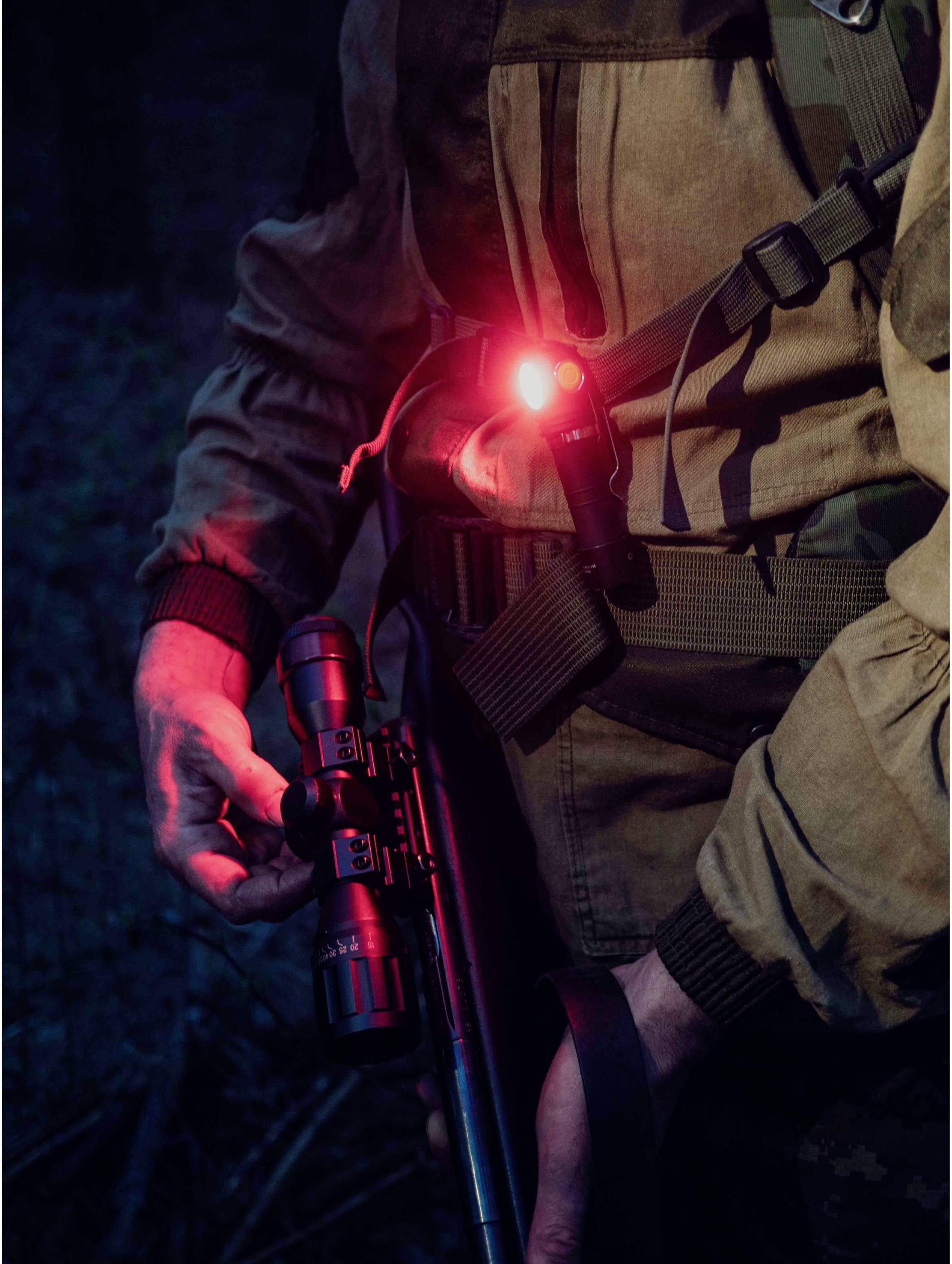 Eine Person in einem dunklen Wald trägt ein Gewehr und ein rotes Licht auf dem linken Arm, das die Umgebung leicht erhellt.