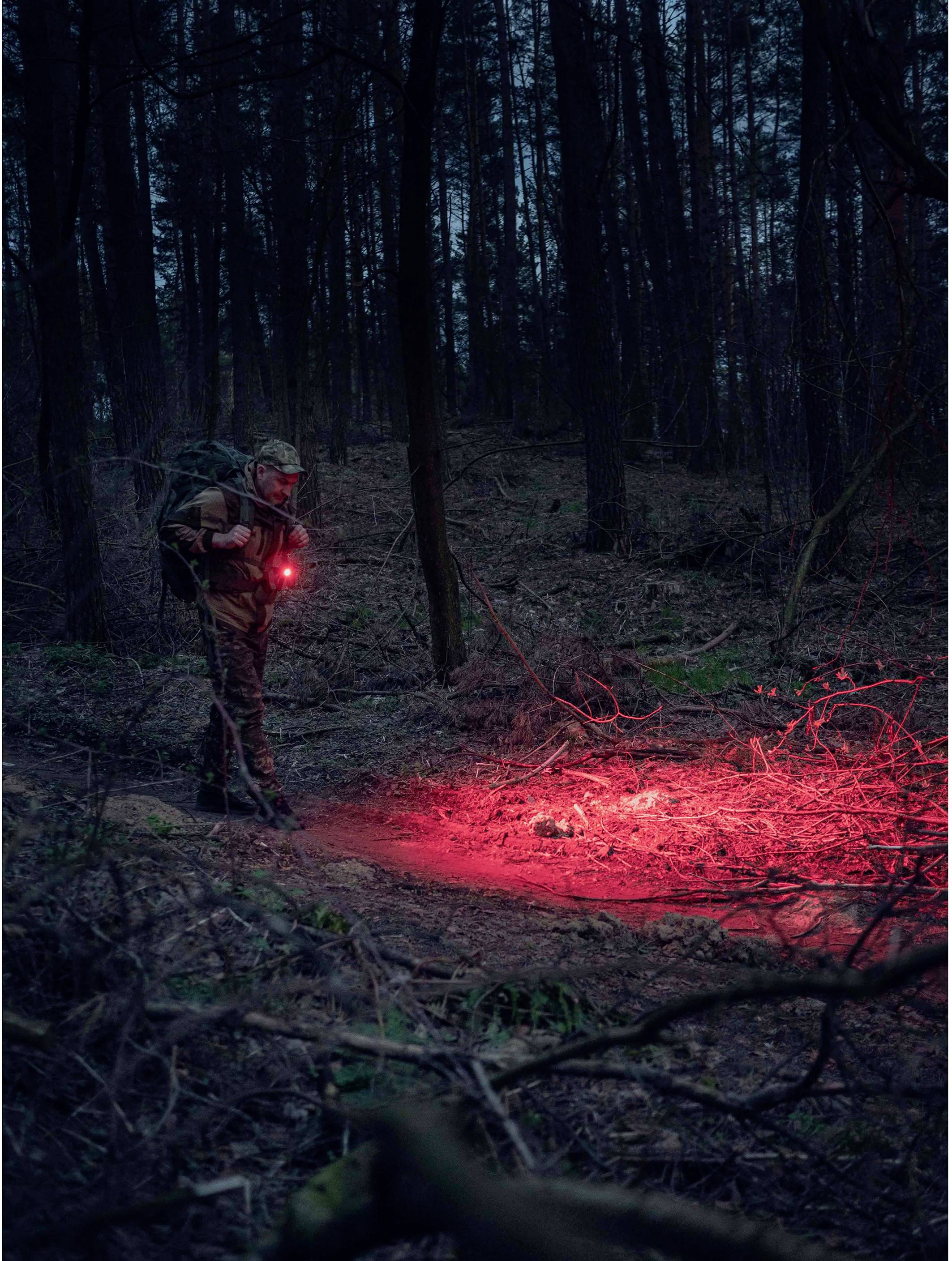Eine Person im Wald bei Dämmerung mit einem roten Licht. Der Boden vor ihr ist ebenfalls rot beleuchtet. Die Umgebung ist dicht bewaldet.