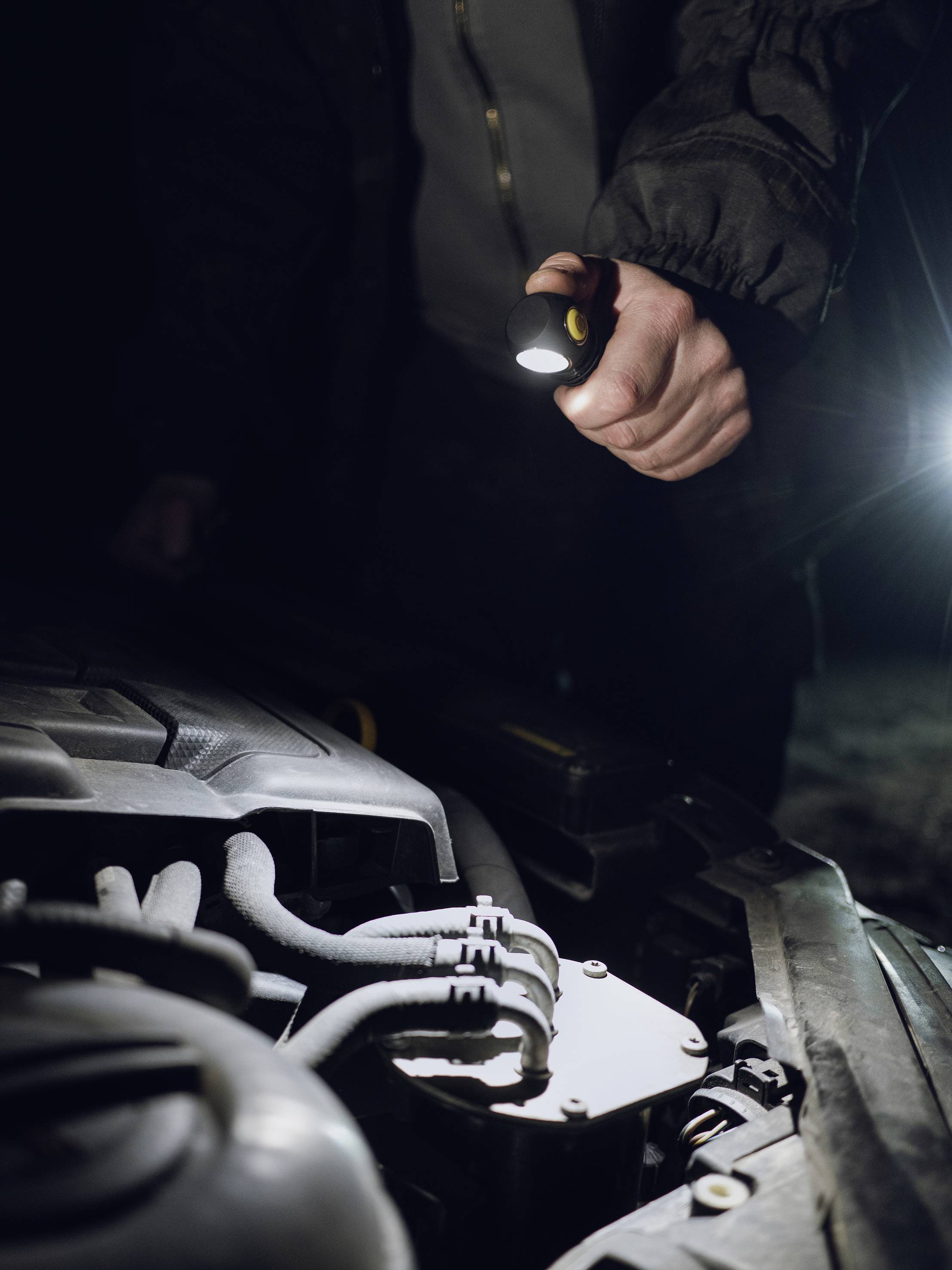 Eine Person beleuchtet mit einer Taschenlampe den Motorraum eines Autos bei Nacht. Die Szene deutet auf eine Fahrzeugreparatur hin.