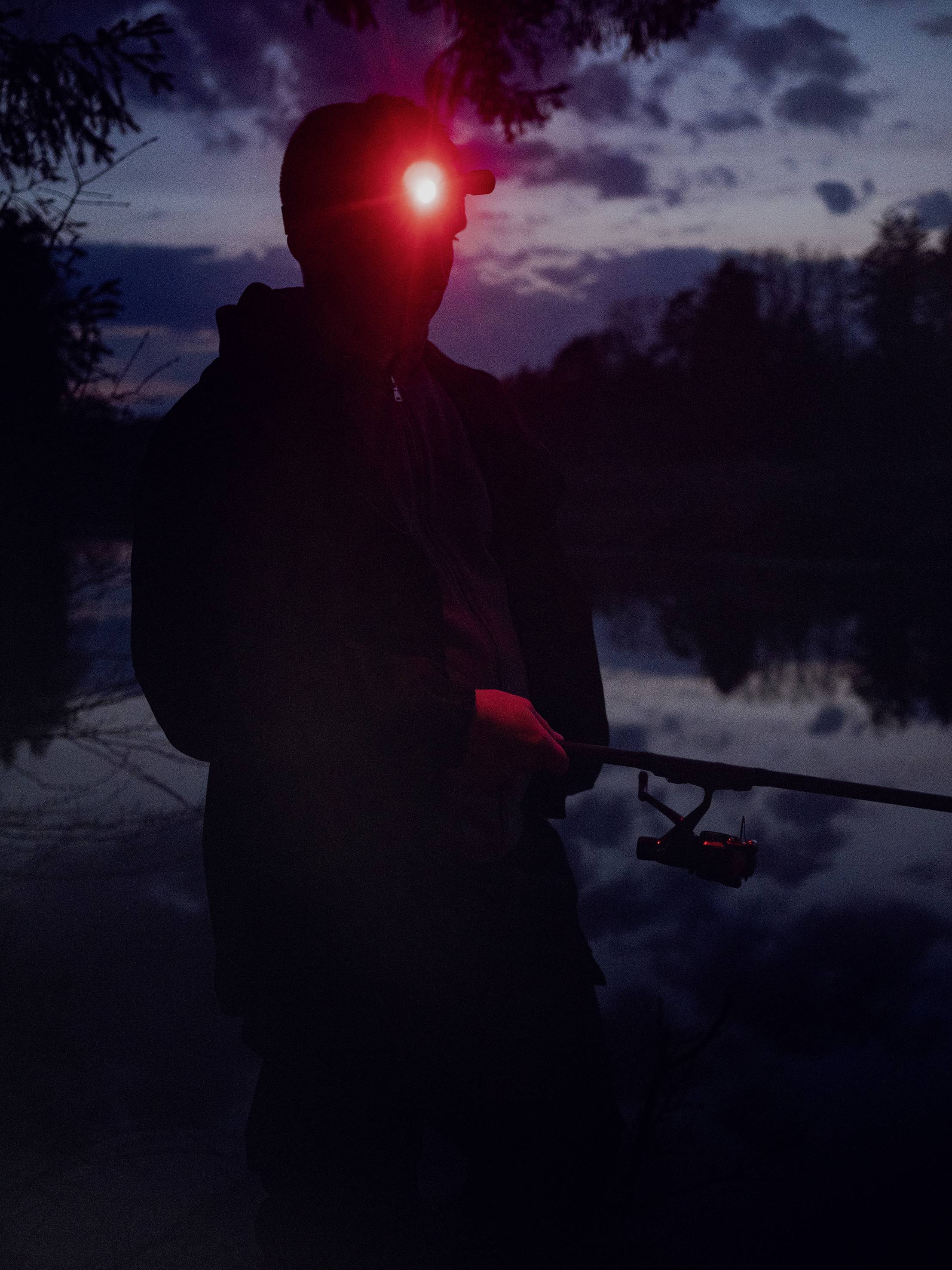 Eine Person angelt im Dunkeln an einem See, beleuchtet von einem roten Stirnlicht. Im Hintergrund ist ein bewaldetes Ufer sichtbar.