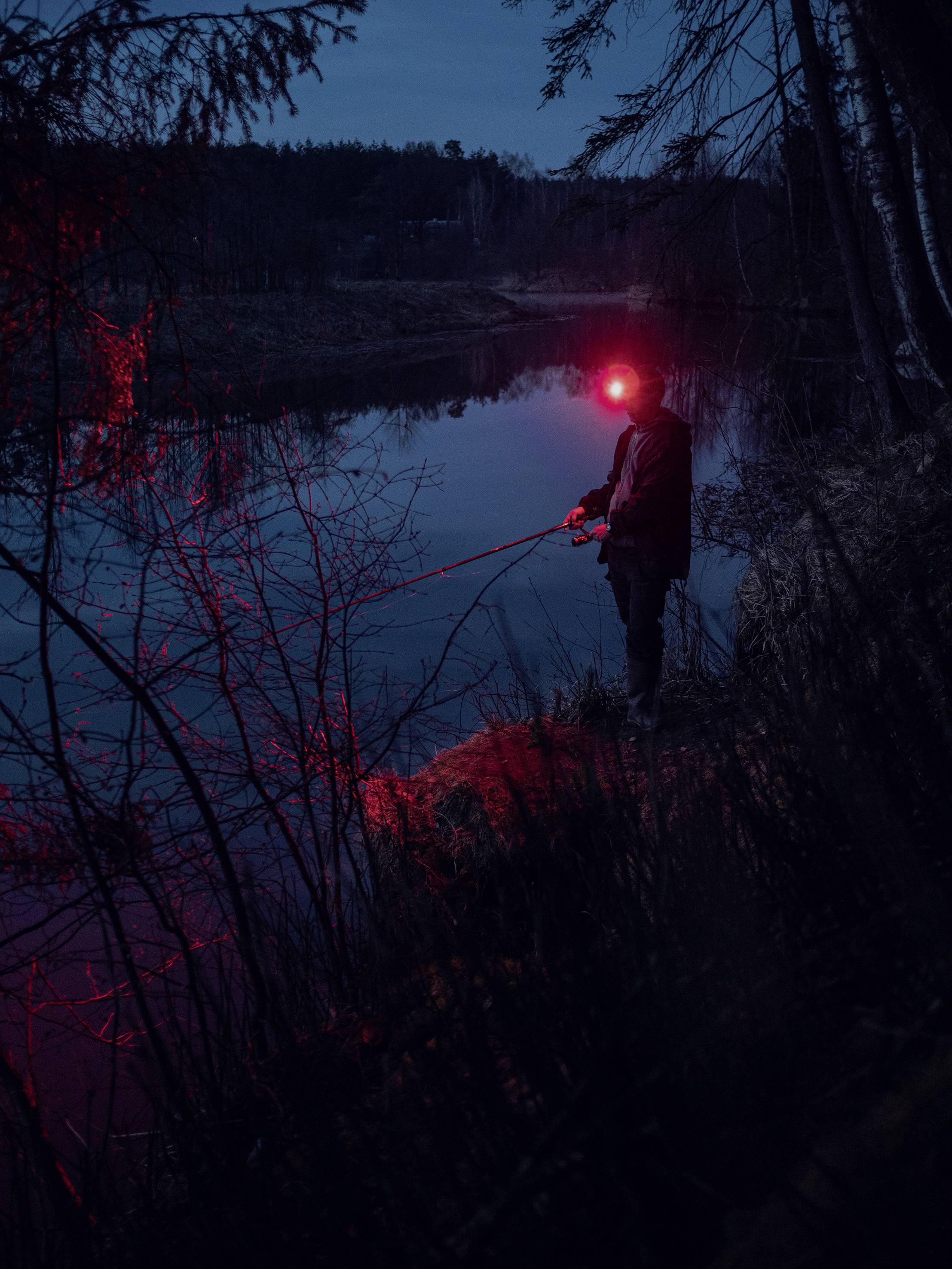 Eine Person angelt nachts am Flussufer, beleuchtet von roten Lichtern. Dunkle Umgebung mit Bäumen und stilles Wasser im Hintergrund.