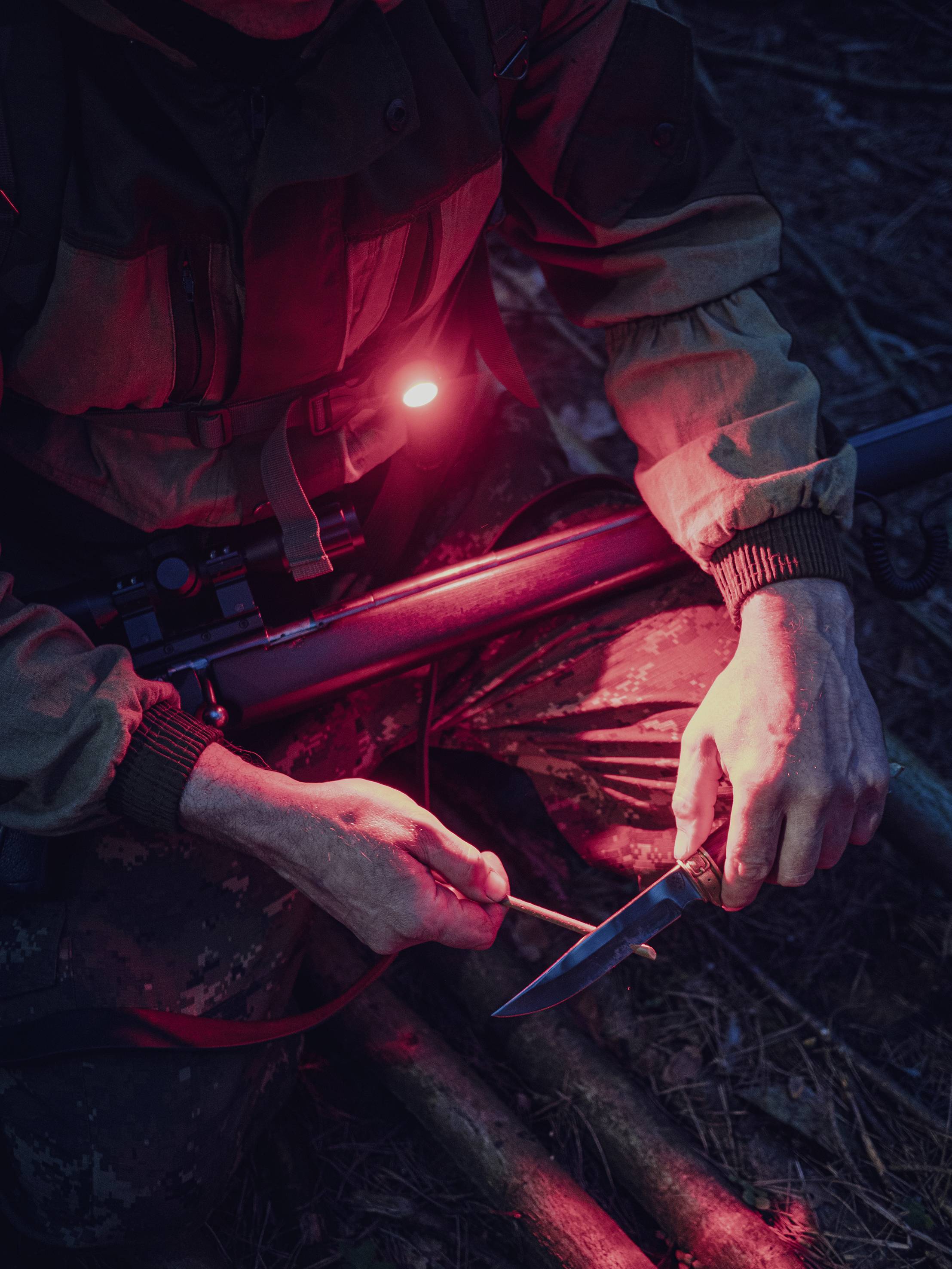 Eine Person sitzt im Wald, schärft ein Messer mit einem roten Licht. Trägt Tarnkleidung, im Freien bei Nacht.