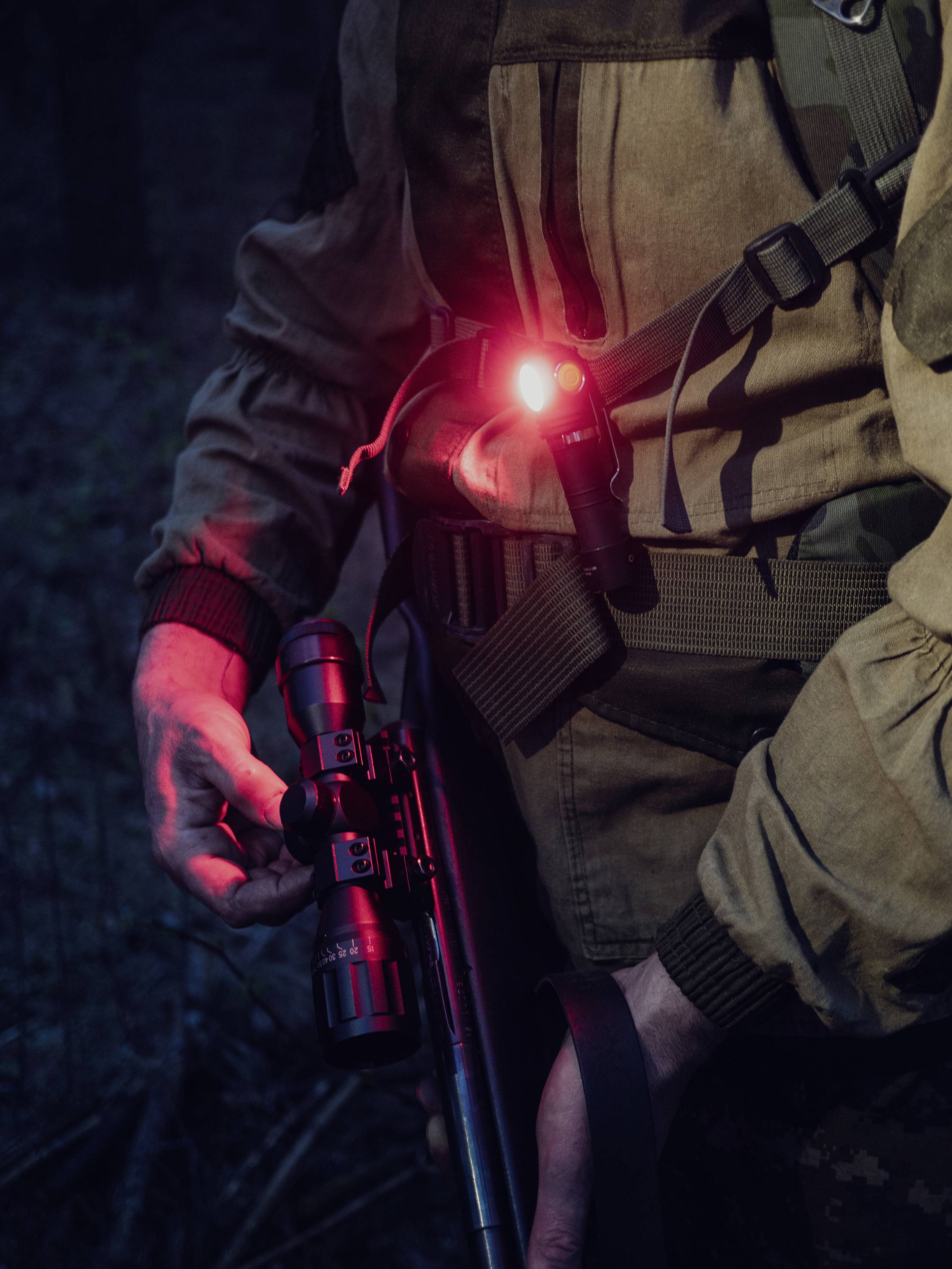 Eine Person hält ein Gewehr und trägt eine olivfarbene Jacke mit einer leuchtend roten Lampe, die ihre Ausrüstung beleuchtet.