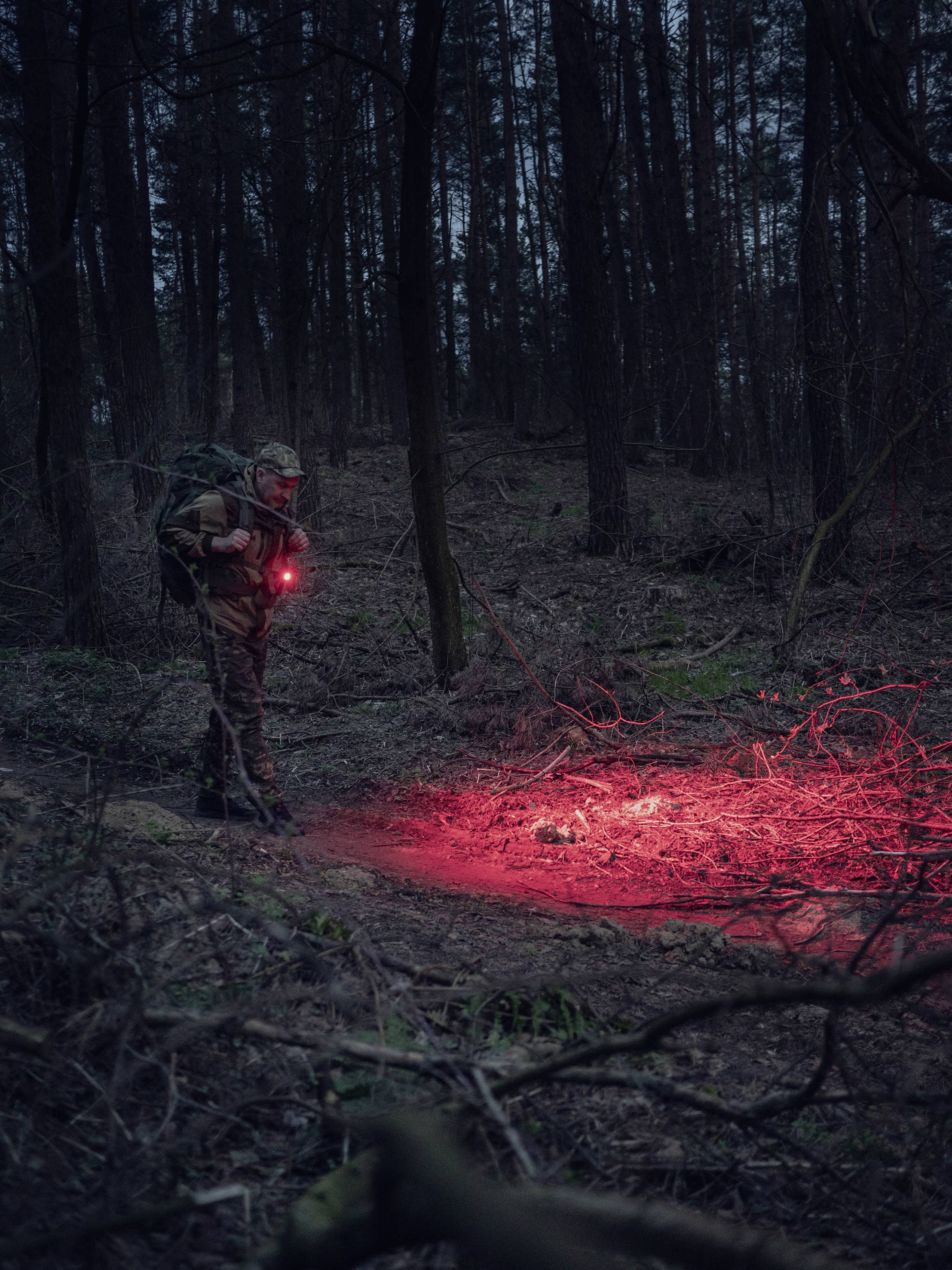 Eine Person in Tarnkleidung steht in einem dunklen Wald und leuchtet mit einer roten Taschenlampe auf einen Bereich vor sich.