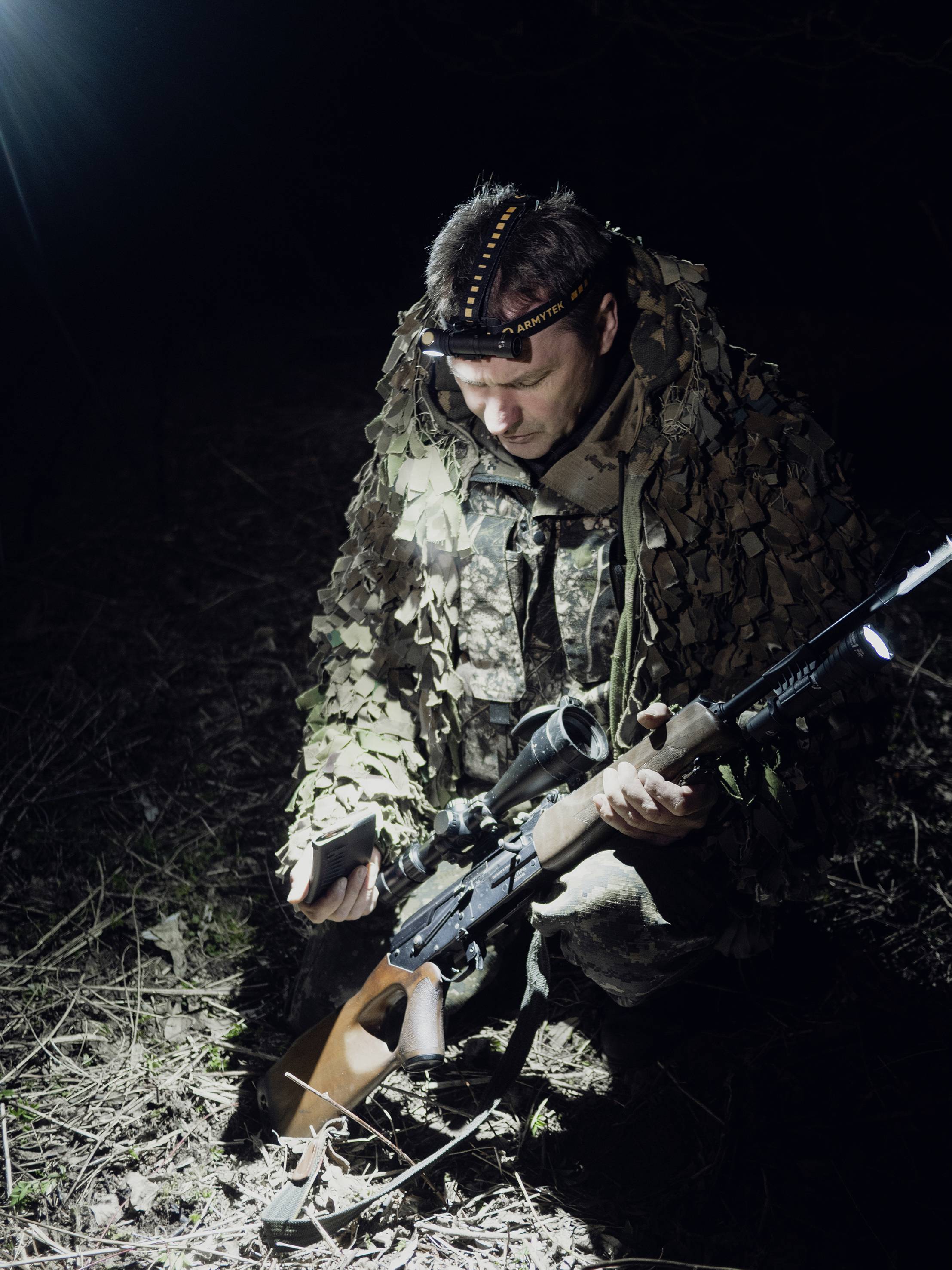 Eine Person in Tarnkleidung kniet im Dunkeln auf dem Boden und hält ein Gewehr. Sie ist von einem intensiven Licht beleuchtet.