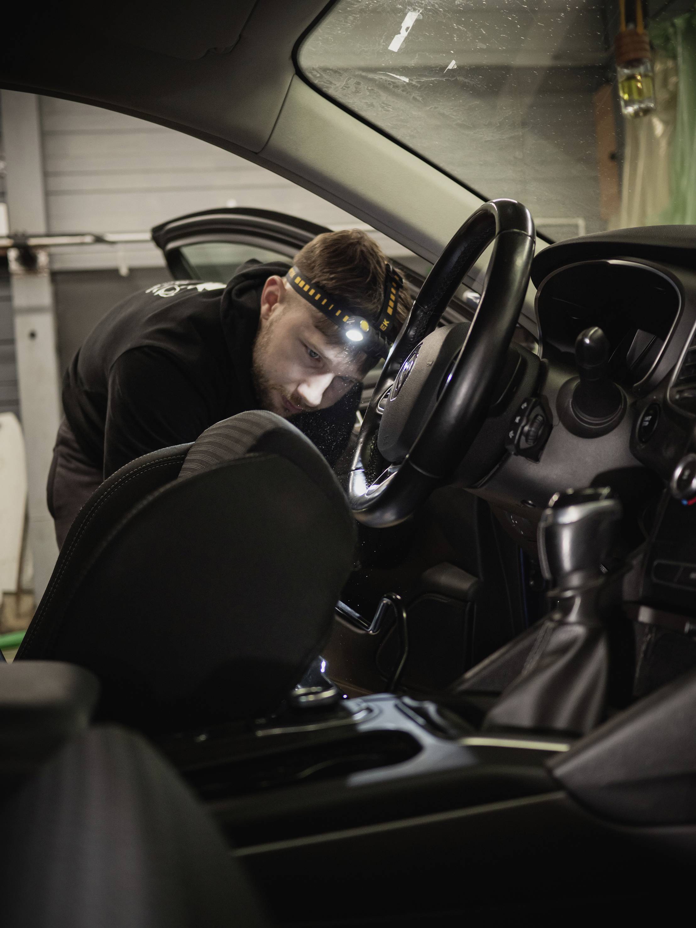 Ein Mechaniker trägt ein Stirnlicht und arbeitet im Inneren eines Autos, konzentriert auf den Bereich des Lenkrads und Armaturenbretts.