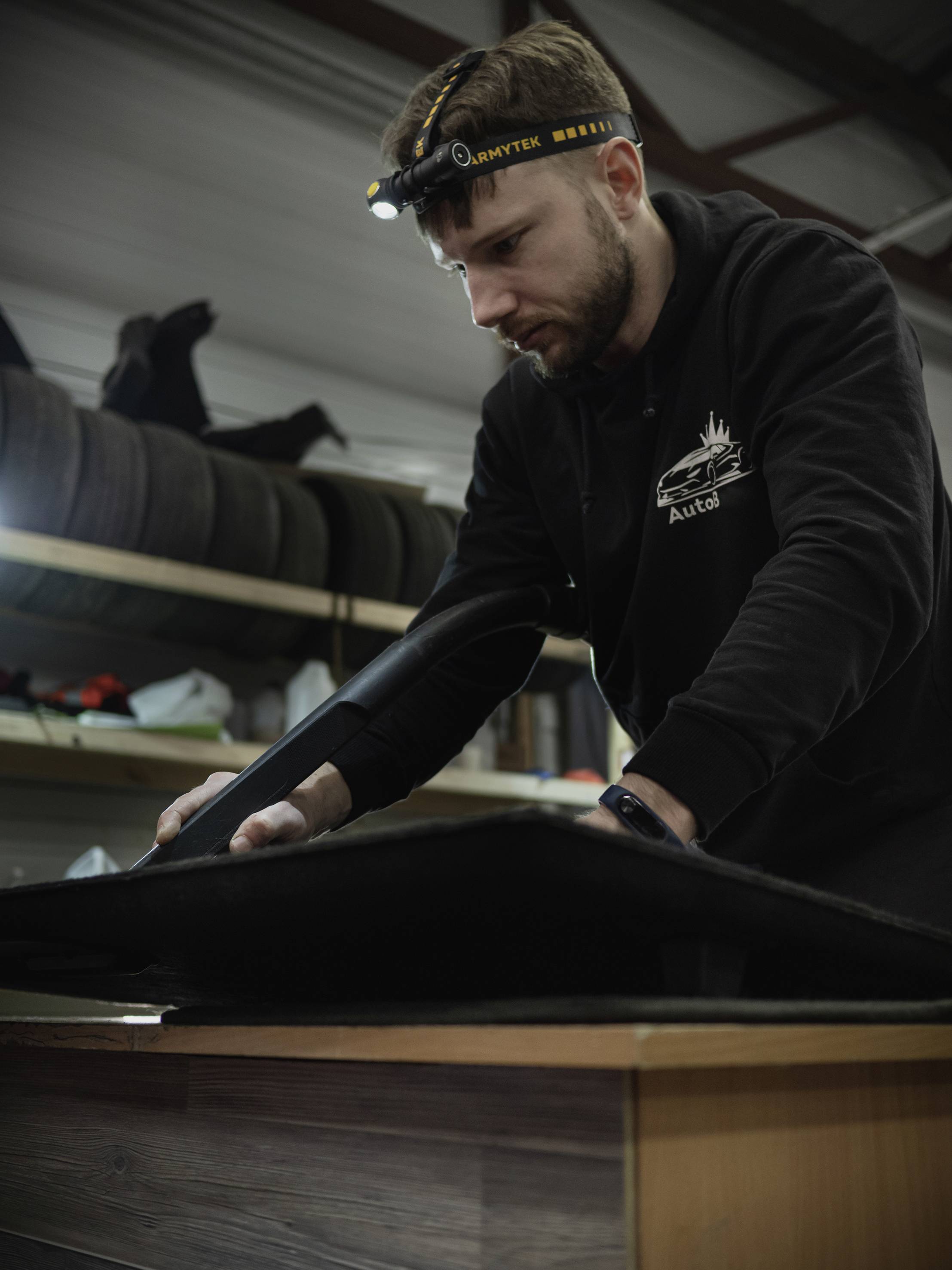 Ein Mann arbeitet konzentriert mit einem schwarzen Tuch in einer Werkstatt, trägt einen Stirn-/Lichtband und einen schwarzen Pullover.