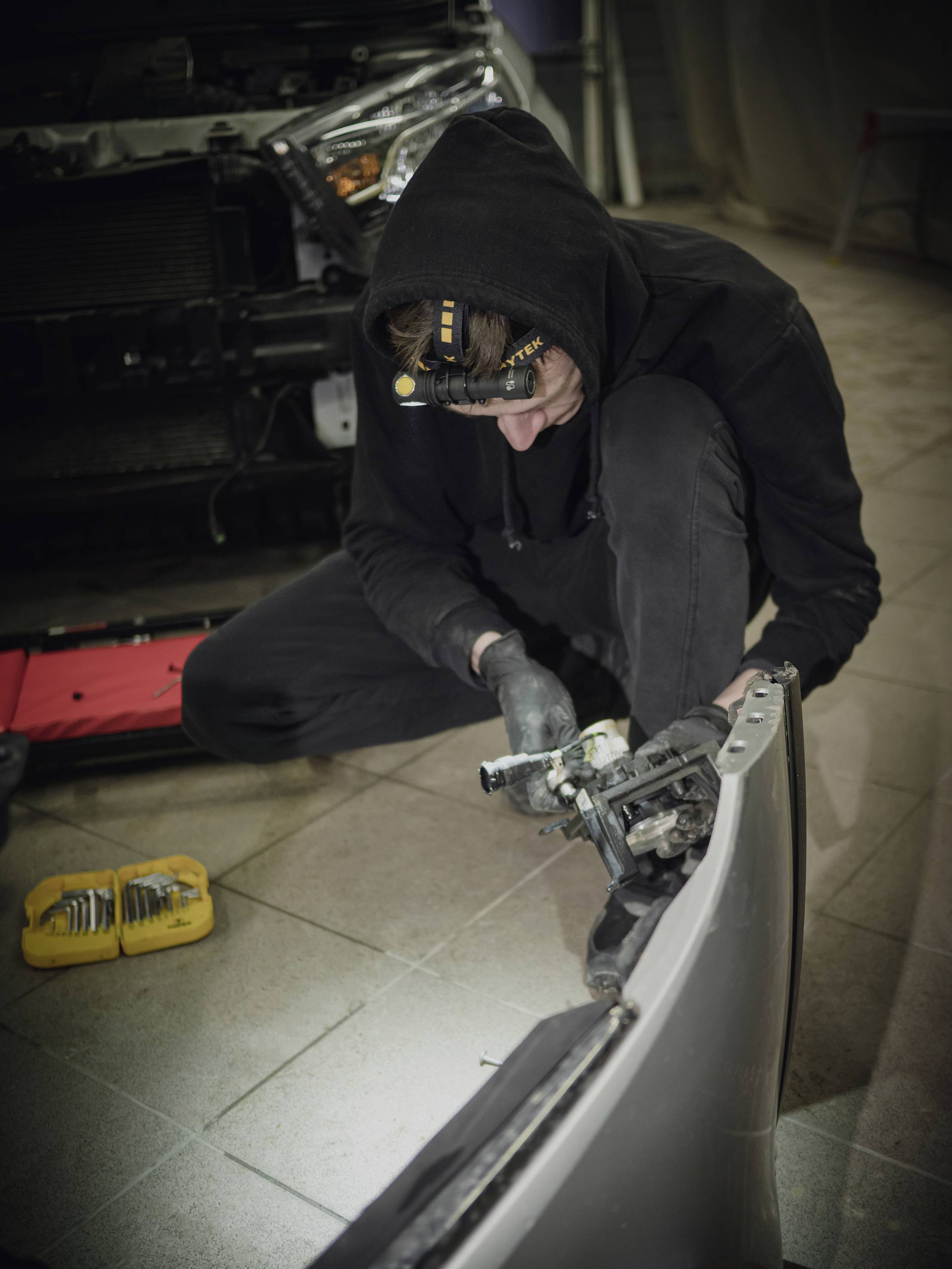 Eine Person in schwarzer Arbeitskleidung repariert ein Autoteil in einer Werkstatt. Ein Auto ist im Hintergrund teilweise zu sehen.
